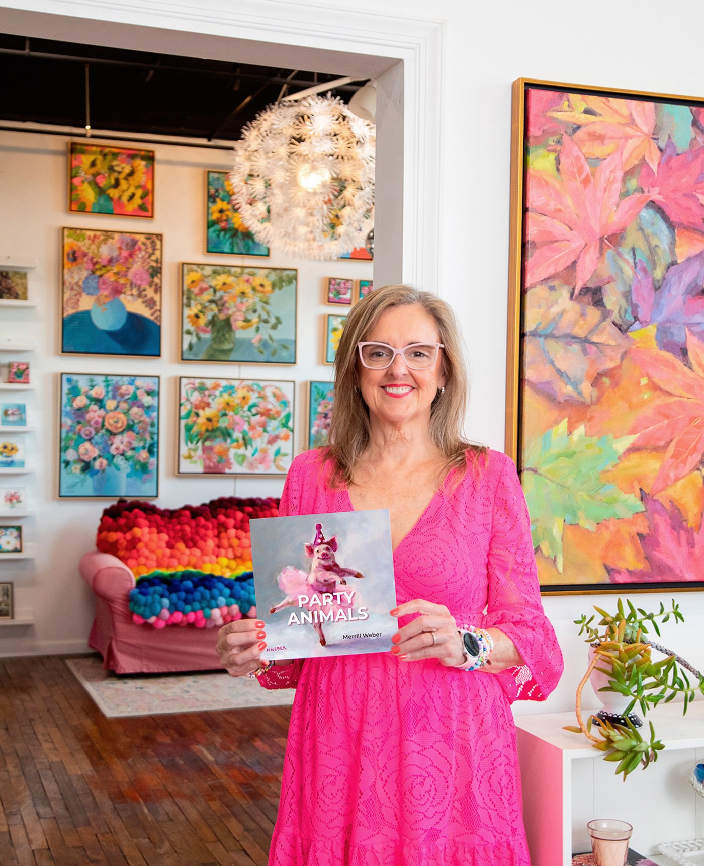 Artist Merrill Weber holding her Party Animals book in her Spring City, Pennsylvania studio and art gallery surrounded by original paintings.