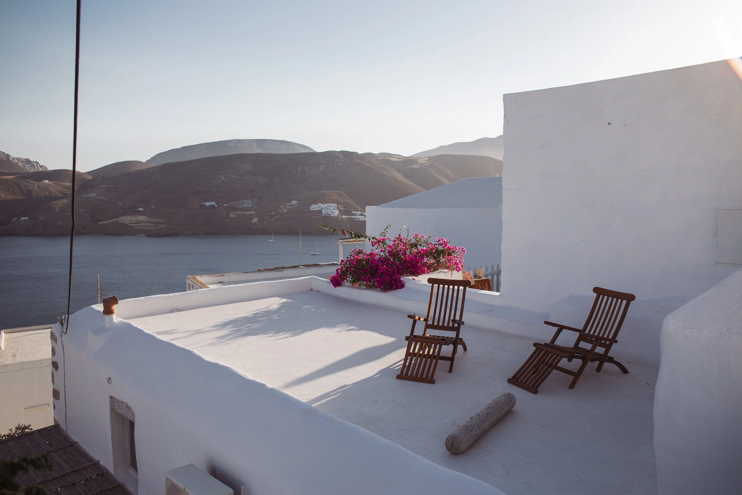 Weiße Gebäude mit Terrasse und Blick auf das Meer und Berge im Hintergrund, rosa Bougainvillea-Pflanze auf Terrasse, zwei Holzstühle, Sonnenlicht
