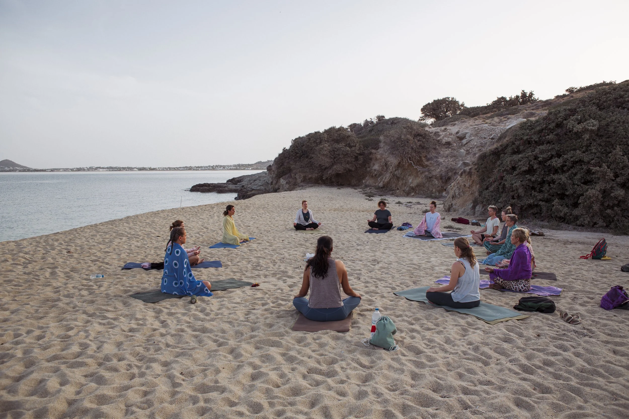Gruppe von Menschen, die auf einem Strand im Kreis Yoga machen, bei Sonnenuntergang, mit Bäumen und Hang im Hintergrund.