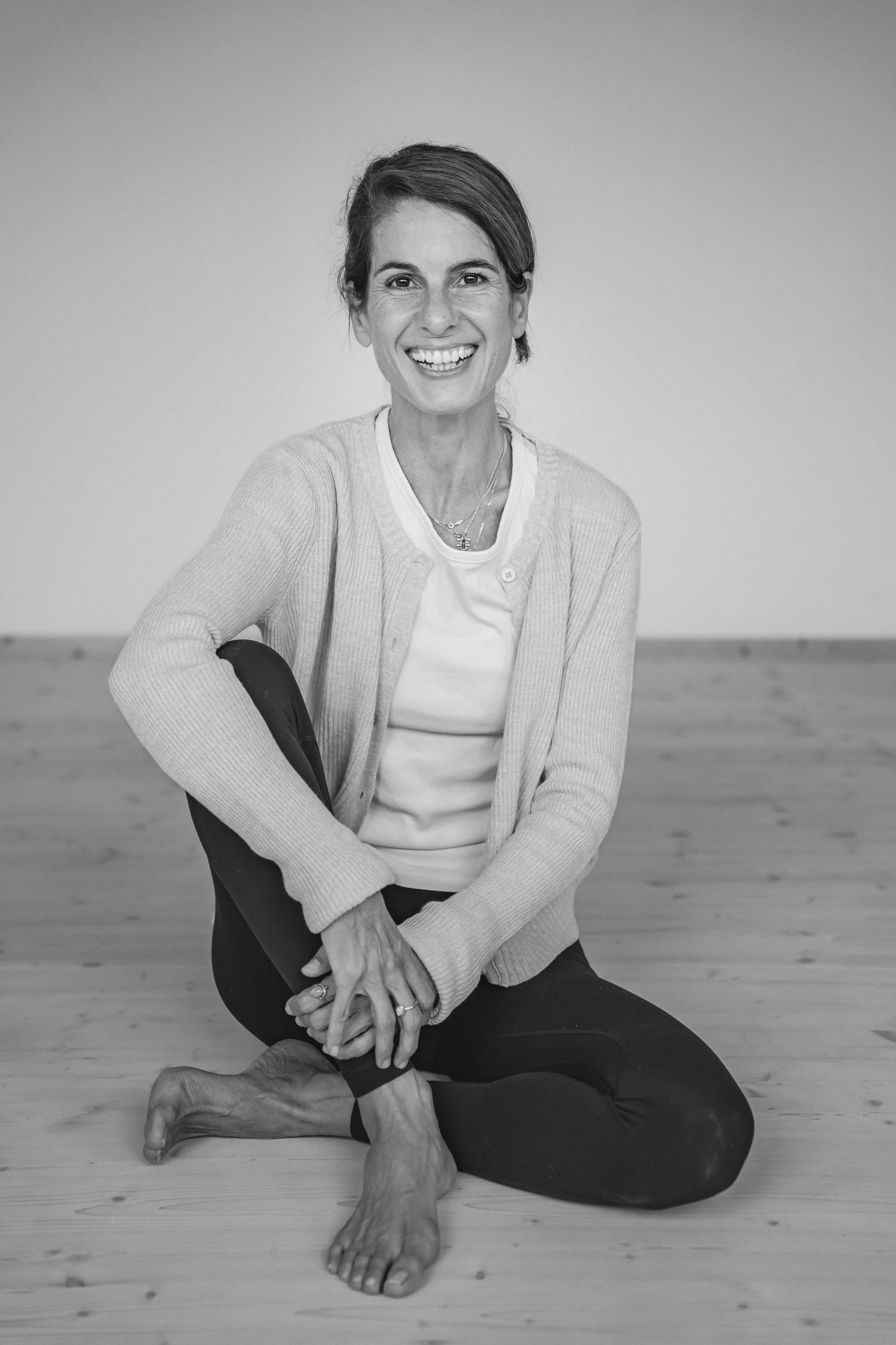 Frau in Yoga-Positur auf Holzboden, lächelnd, in einem Raum mit hellem Hintergrund, schwarze Leggings, graue Strickjacke, weiße T-Shirt, barfuß.