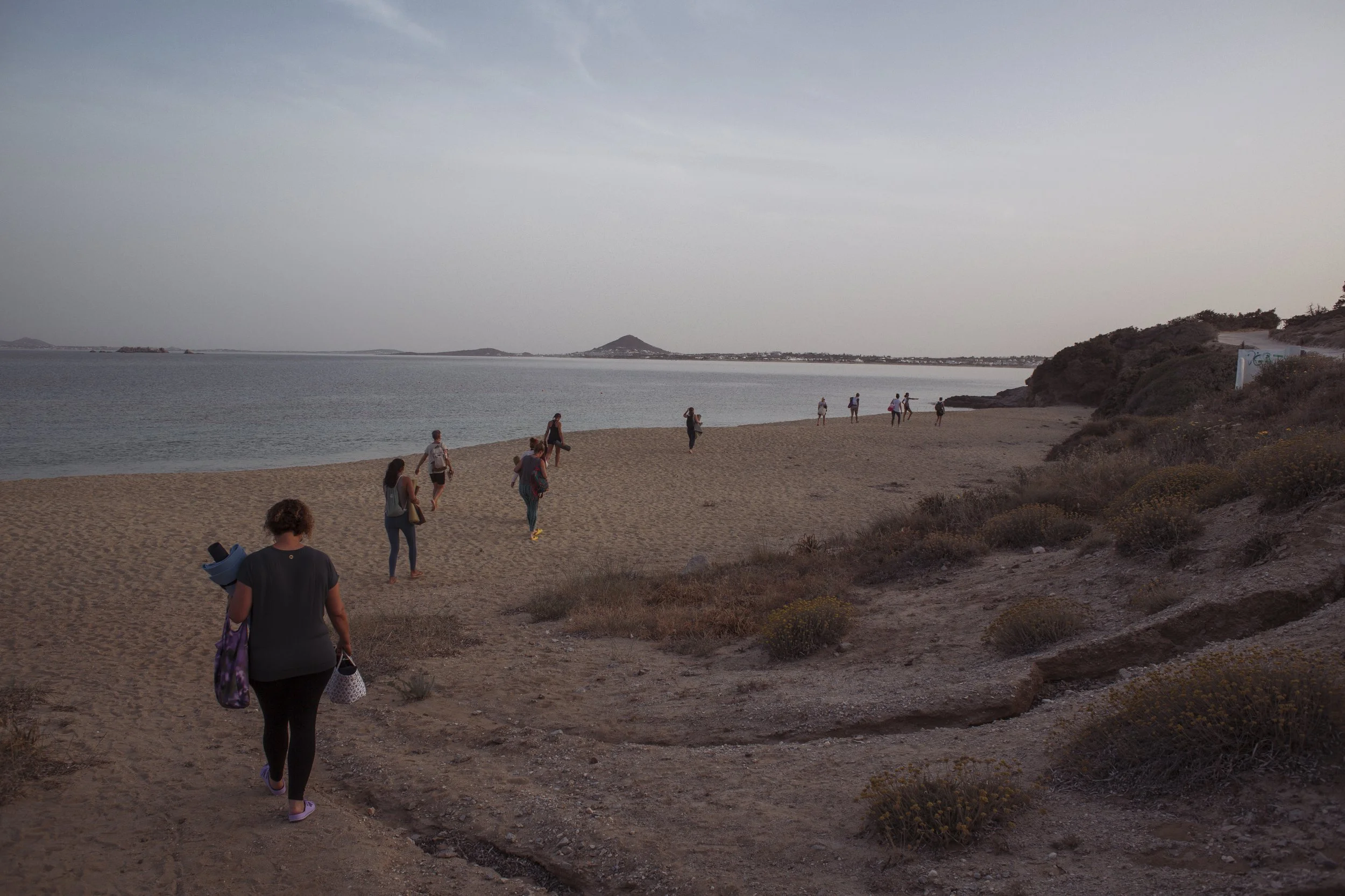 Menschen gehen an einem Strand entlang, während die Sonne untergeht, mit Blick auf das Wasser und entfernte Inseln im Hintergrund.