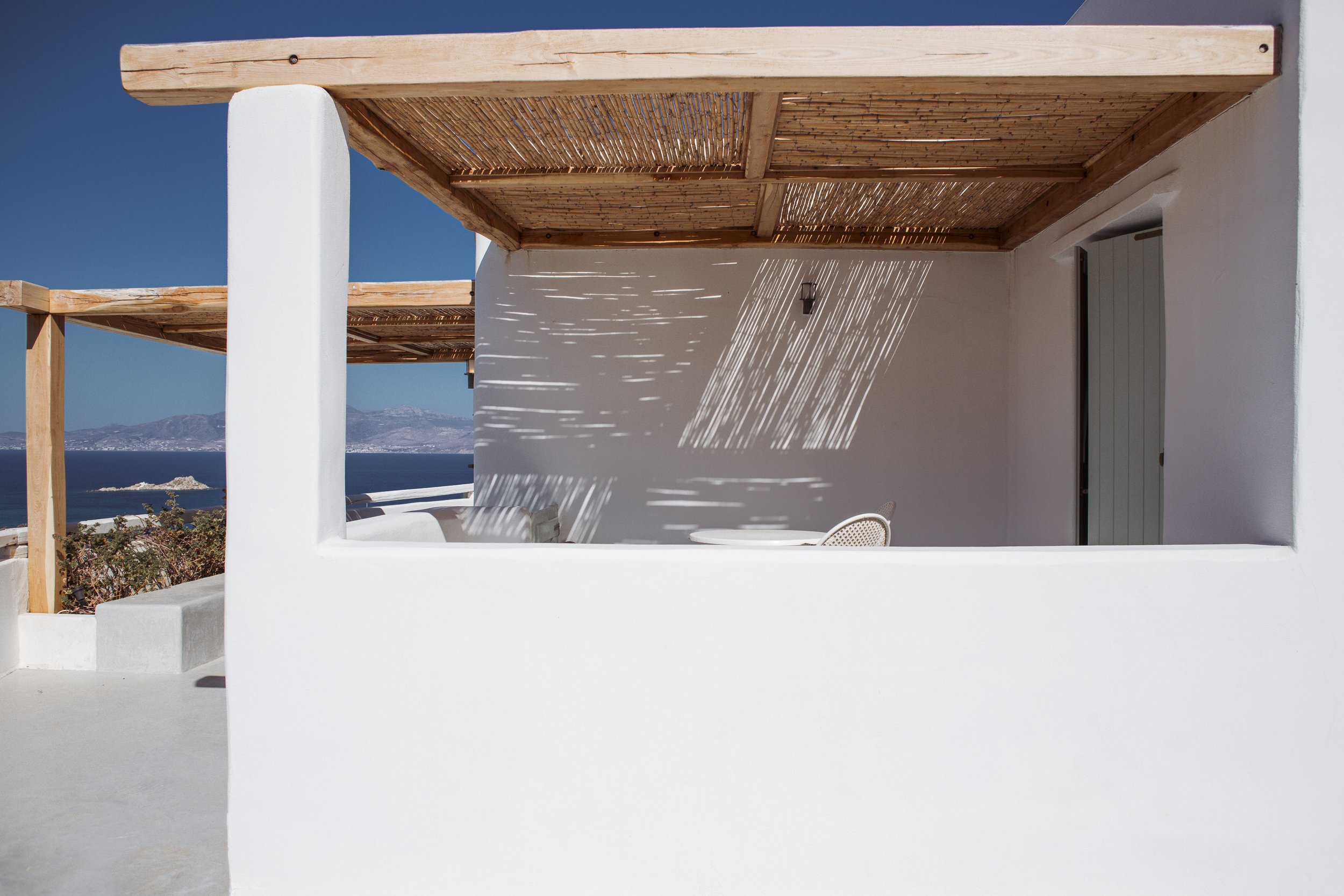 Blick auf eine weiße Terrasse mit Rattanmöbeln unter einem Holzdach, mit Blick auf das blaue Meer und Berge im Hintergrund, Schatten von Lamellen auf die Wand projiziert.