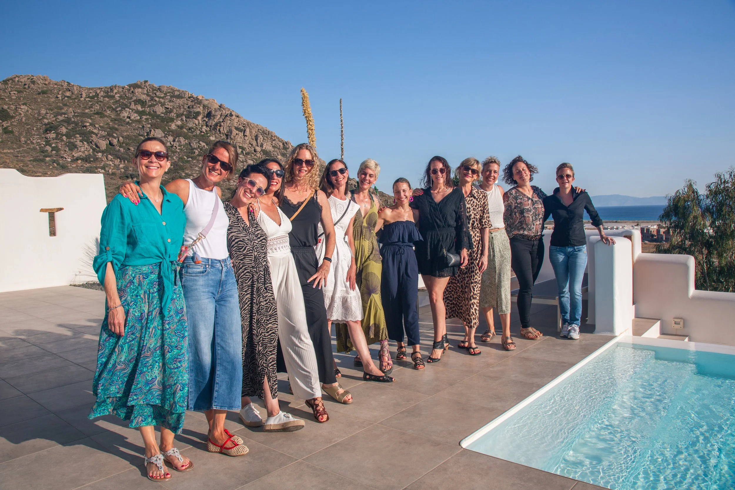 Eine Gruppe von Frauen steht nahe einem Pool im Freien mit Blick auf Berge und Wasser, genießt die Sonne und trägt sommerliche Kleidung.