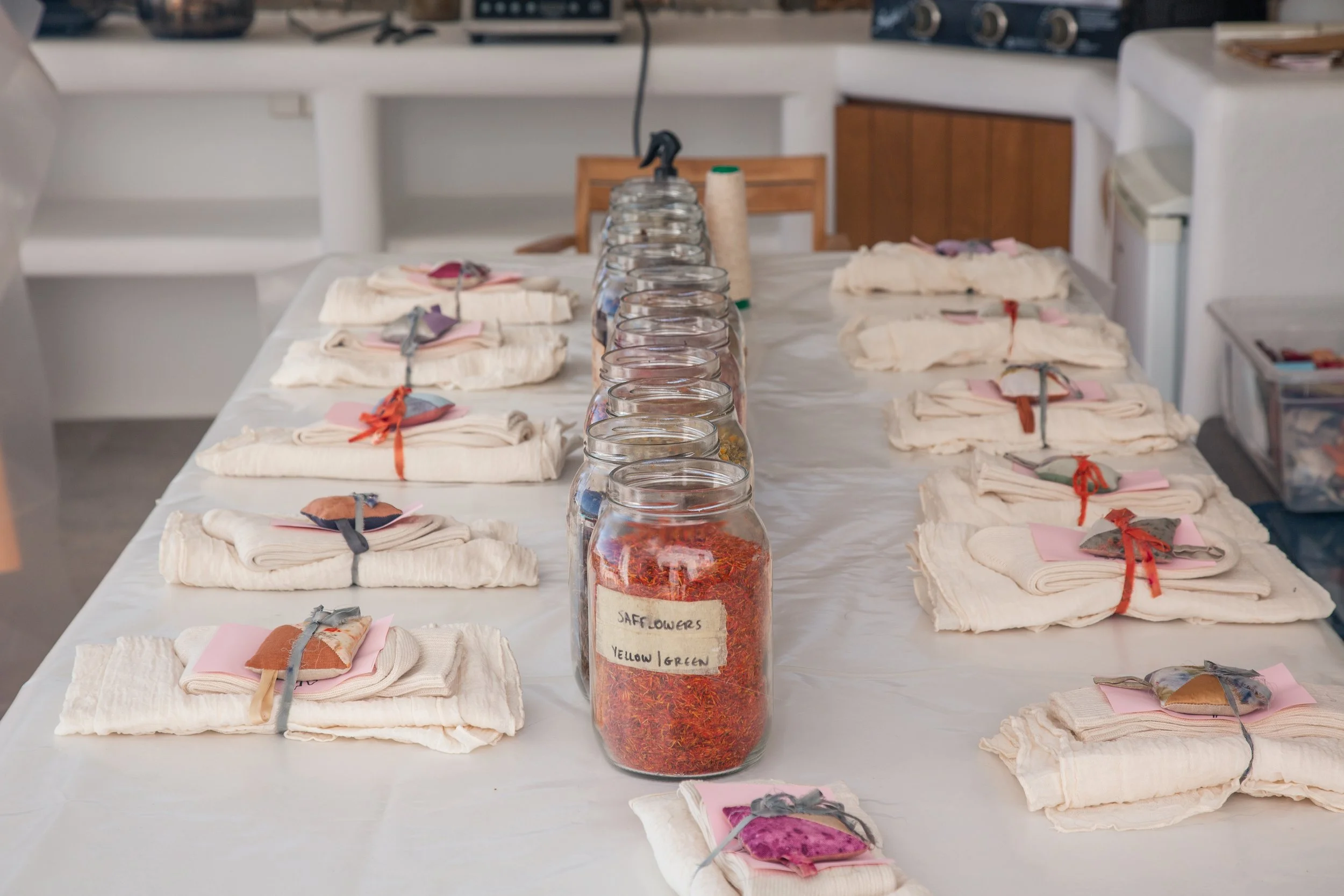 Bunte, festlich verzierte Stoffstückchen, die auf weißen Tüchern liegen, neben am Tisch in Gläsern aufbewahrtem getrocknetem Blumenmaterial, vermutlich für die Herstellung von Floristik- oder Bastelarbeiten.