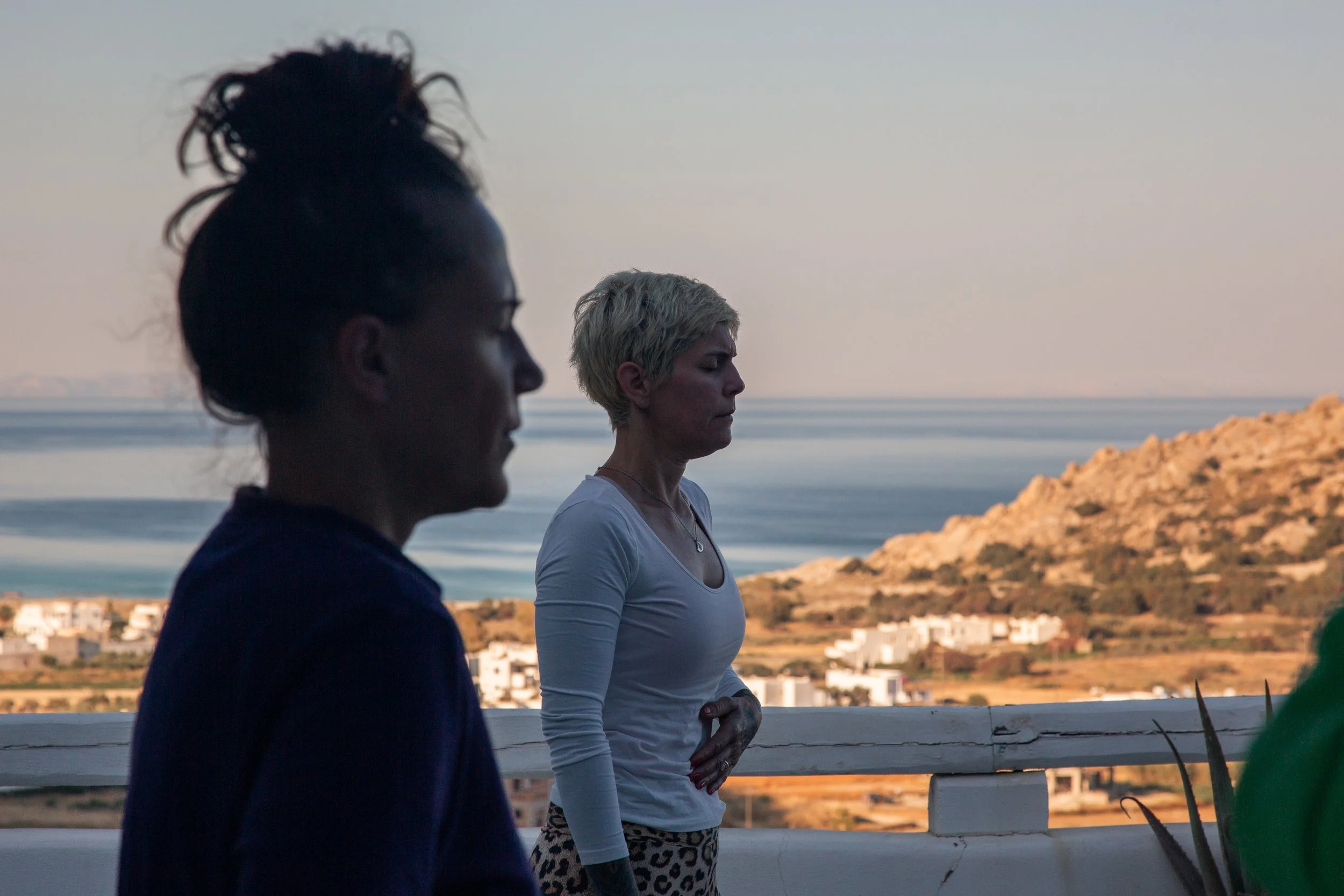 Zwei Frauen stehen auf einer Terrasse mit Blick auf das Meer und eine bergige Landschaft bei Sonnenuntergang, eine neben der anderen, eine in Profil, die andere mit Blick nach vorne.