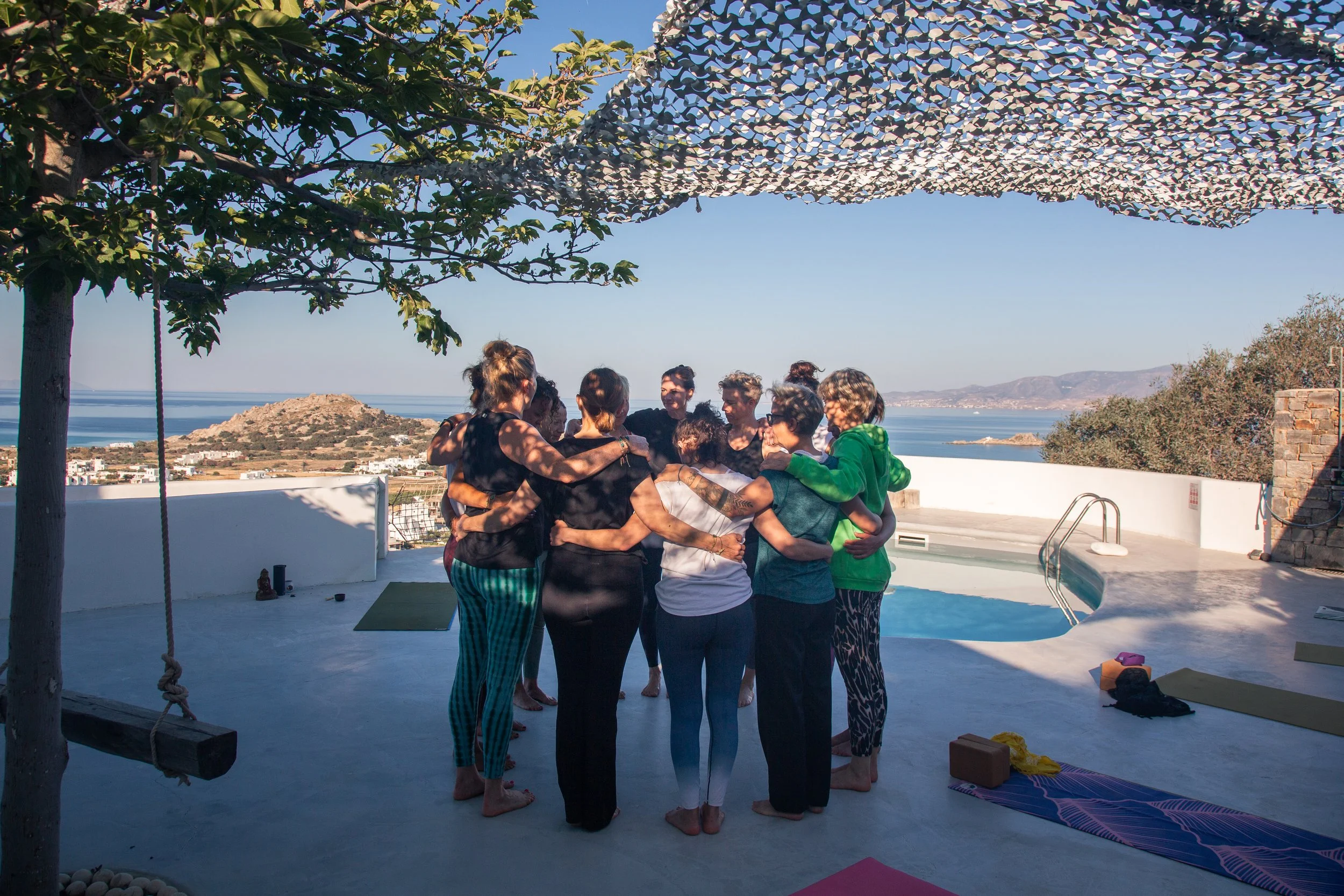 Gruppe von Menschen in einer Huddle-Position im Freien mit Blick auf das Meer, umgeben von Vögeln, unter einem schattigen Netz, bereit für eine Yoga- oder Meditationssitzung
