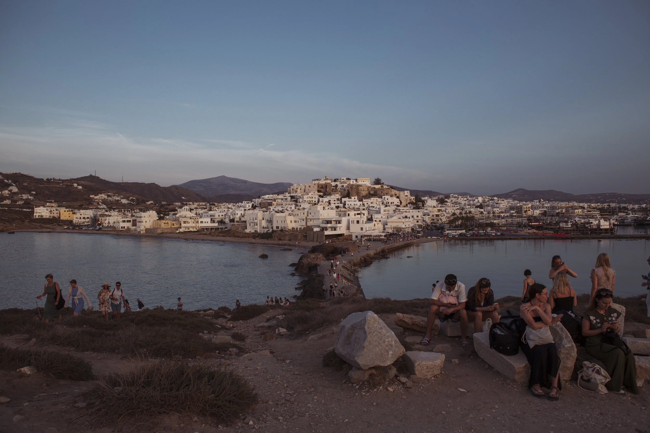 Blick auf ein Küstenstädtchen mit weißen Gebäuden auf einer Anhöhe, Menschen sitzen und gehen auf dem Weg am Wasser entlang, im Vordergrund sitzen Personen, die auf Steinen sitzen oder unterwegs sind, bei Sonnenuntergang oder Dämmerung.