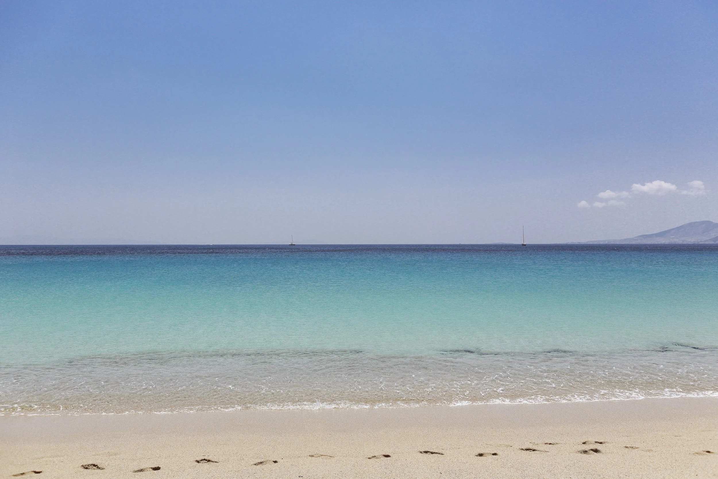 Ein Strand mit goldenem Sand, blauem Wasser und einem klaren Himmel, mit zwei Segelbooten im Meer.