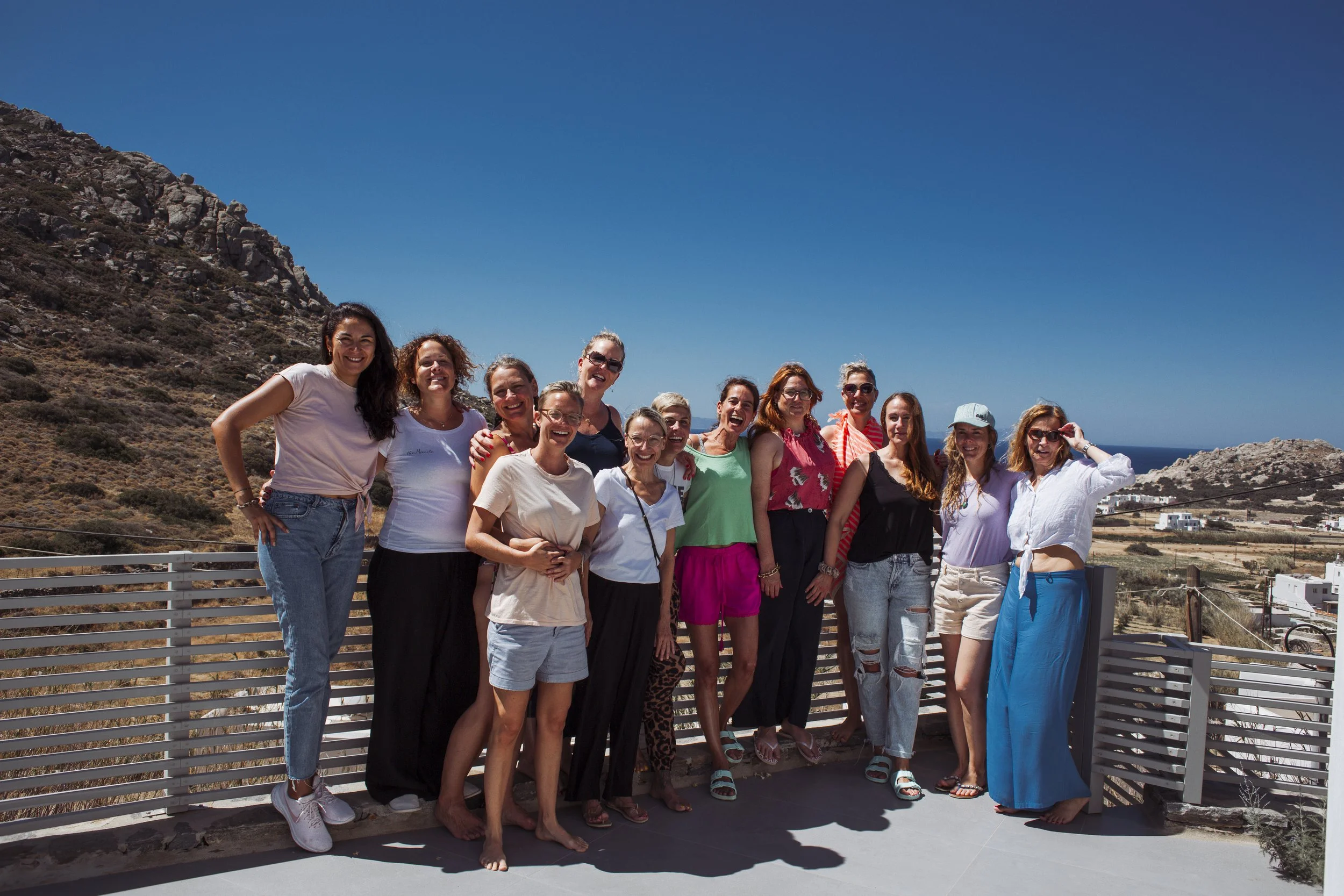Gruppe von 14 Frauen, die auf einer Terrasse im Freien vor einer Berglandschaft und dem Meer posieren, alle lachen und tragen sommerliche Kleidung.