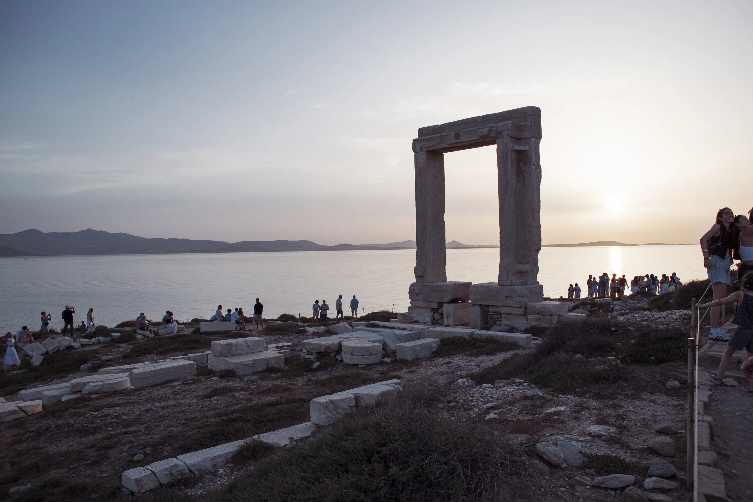 Alter Torbogen im Sonnenuntergang am Meer, mit vielen Touristen und Spaziergängern im Hintergrund.