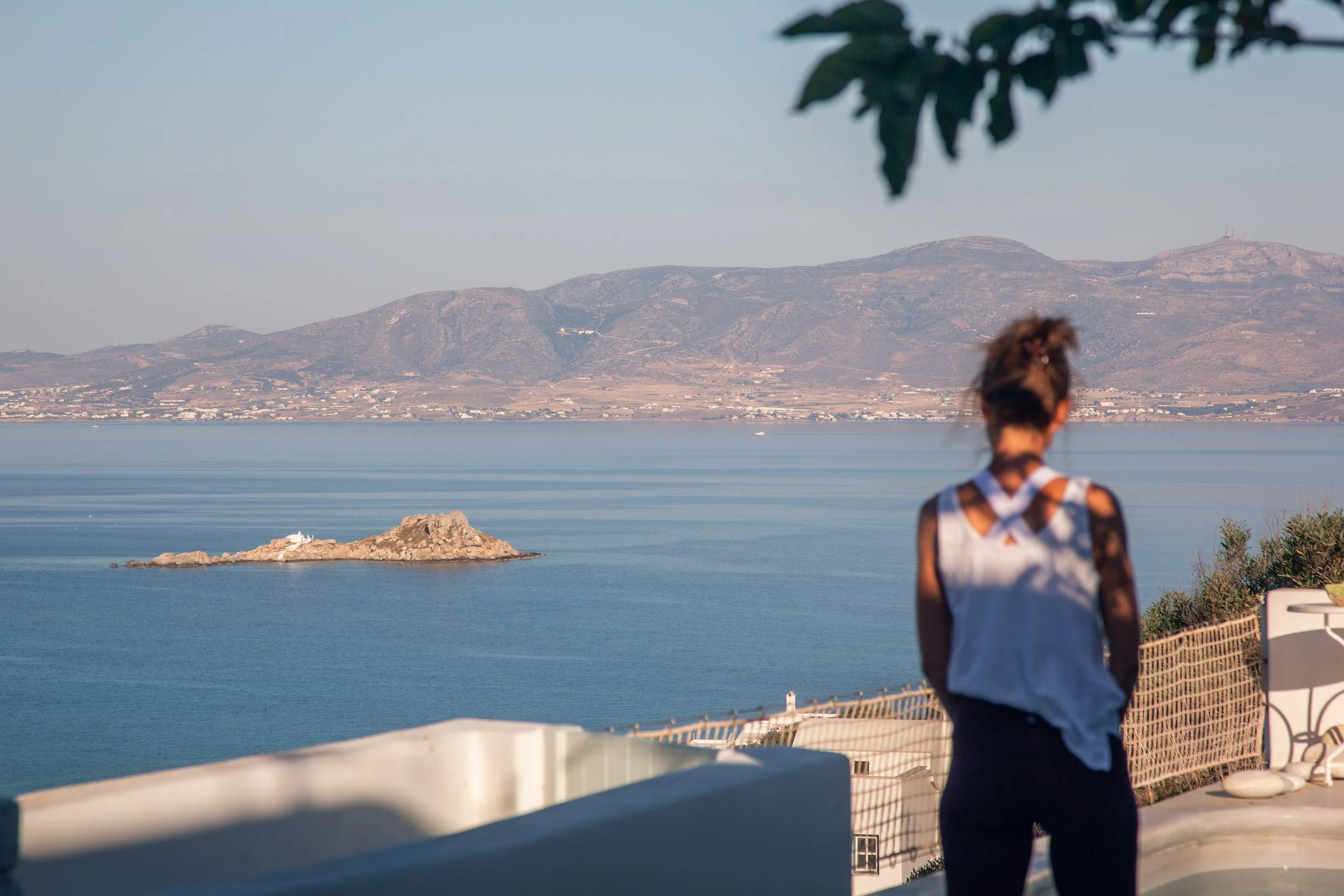 Frau blickt auf das Meer und die Insel mit Leuchtturm im Vordergrund, Berge im Hintergrund, sonniger Tag.
