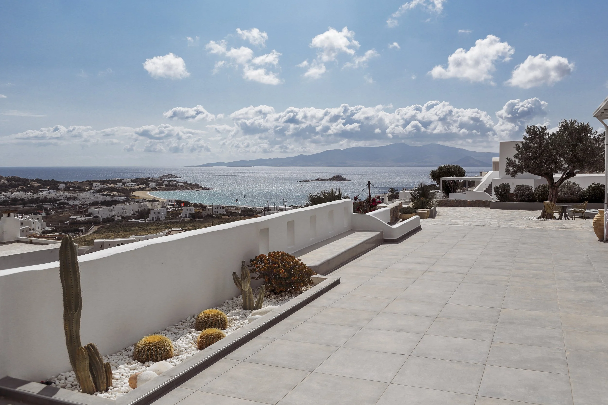 Blick auf den Meereshorizont mit weißen Gebäuden an der Küste und einem Berg im Hintergrund, davor eine Terrasse mit weißer Mauer, Pflanzen und Stühlen.