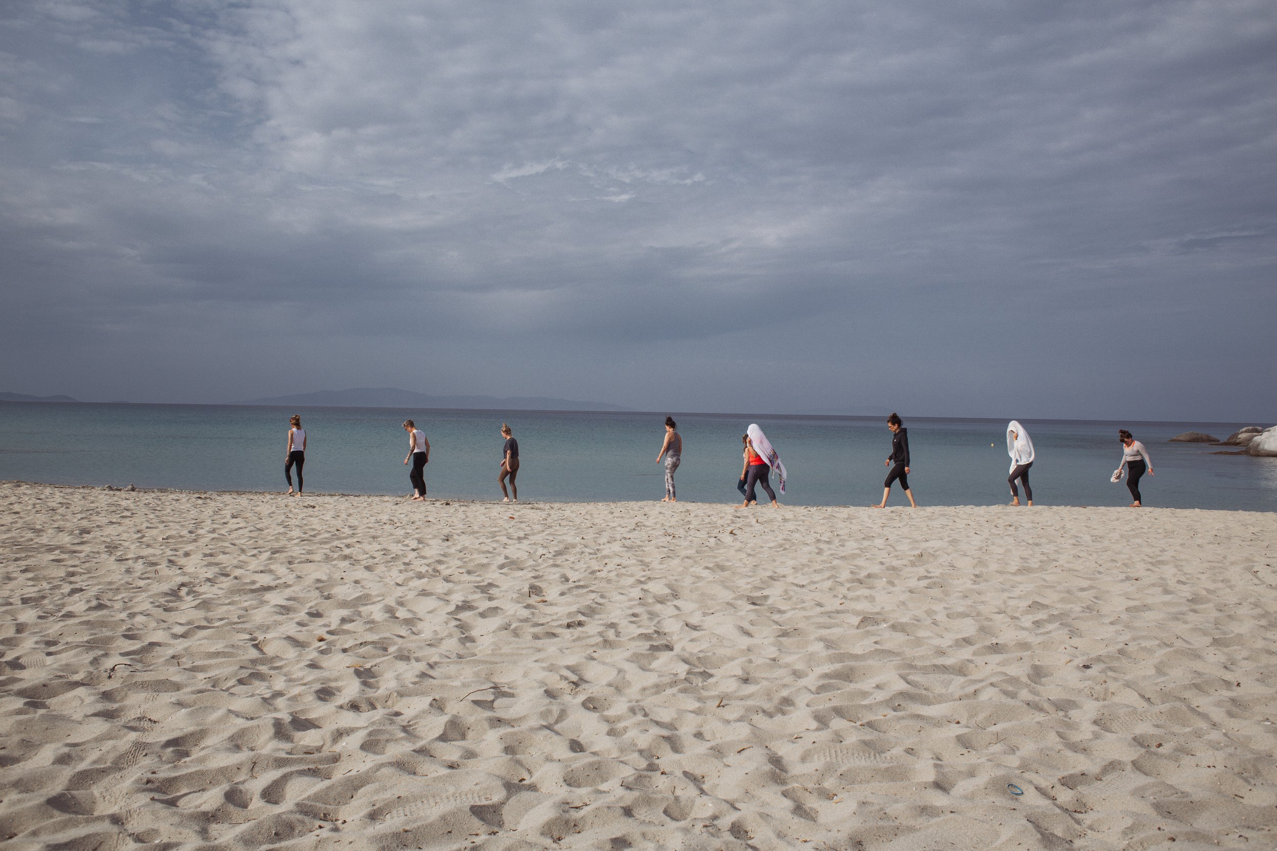 Mehrere Frauen spazieren am Strand entlang, einige tragen Kopftücher, im Hintergrund das ruhige Meer und ein bewölkter Himmel.