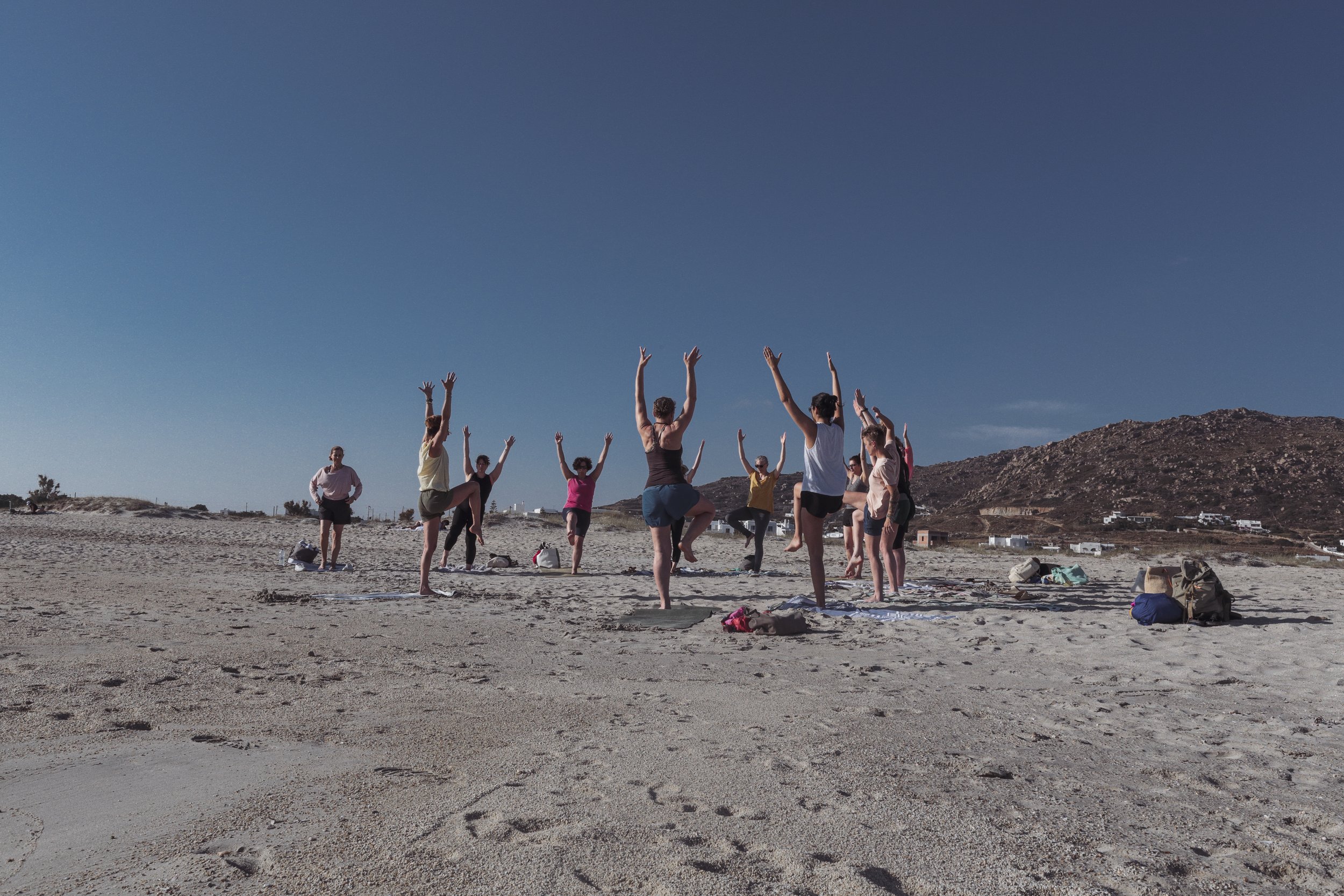 Gruppe von Menschen beim Yoga am Strand unter blauem Himmel, umgeben von Sand und Bergen im Hintergrund.