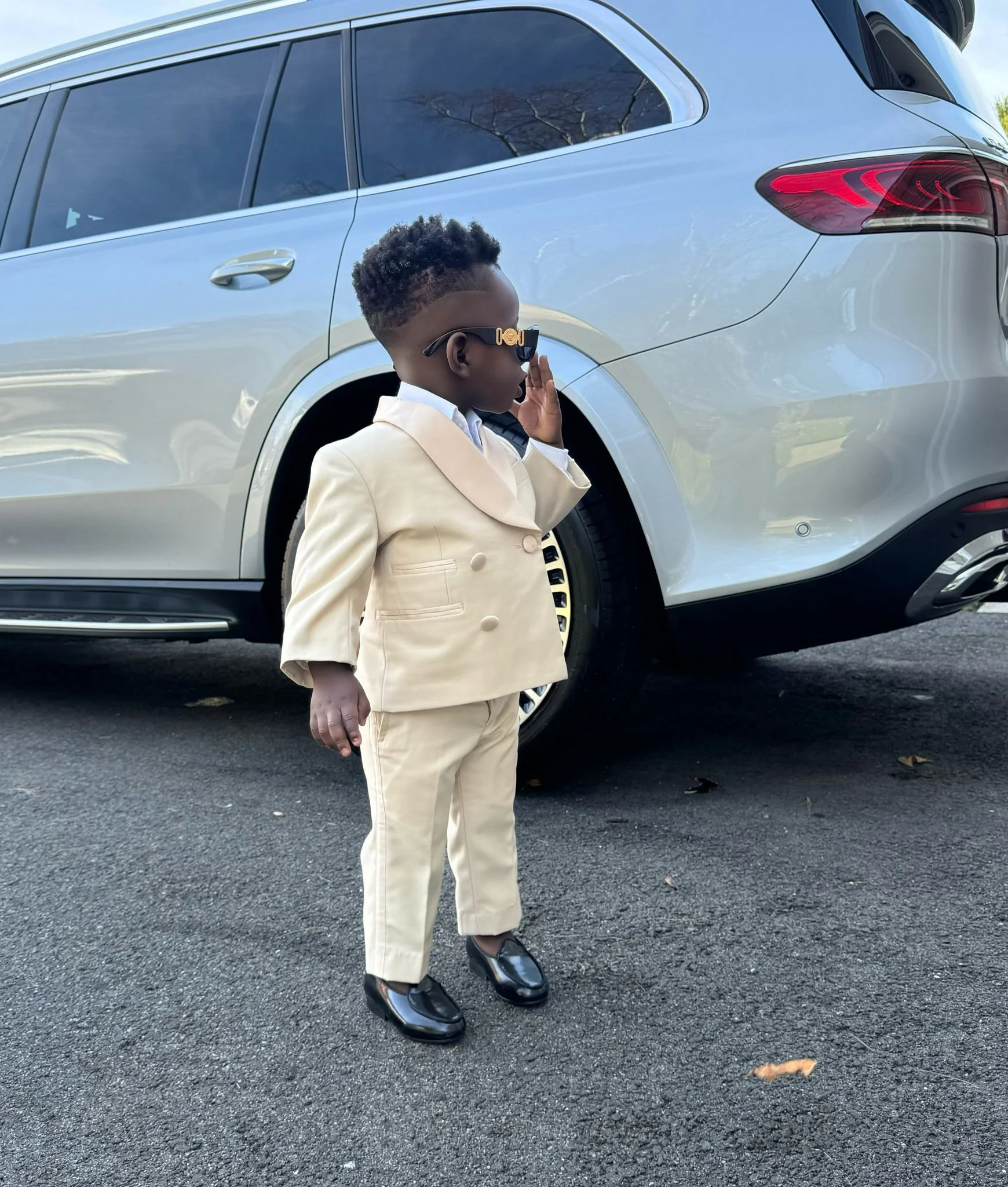 A young boy dressed in a beige suit, black shoes, and sunglasses, standing on a paved area near a silver car, saluting with one hand.
