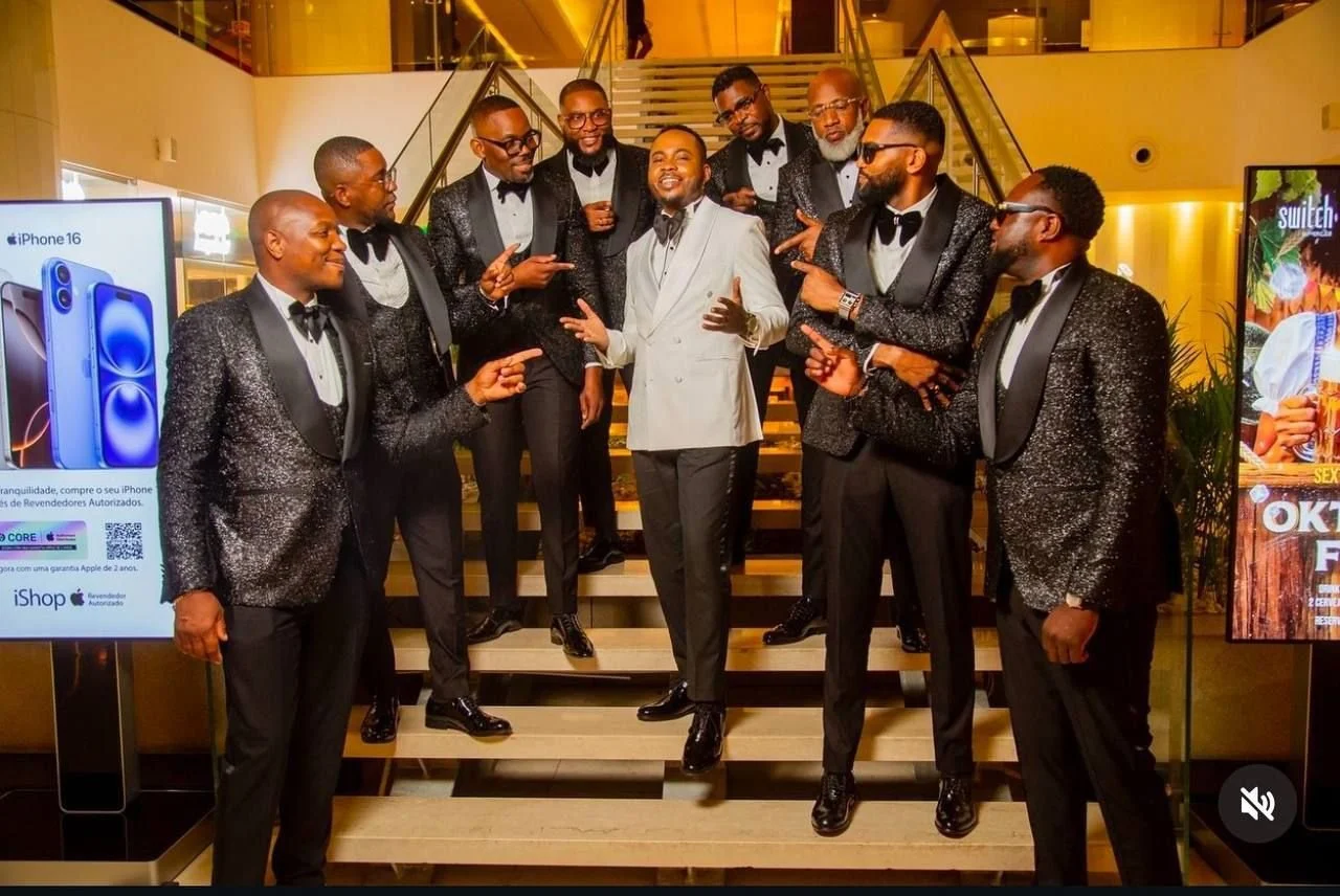A group of ten men dressed in tuxedos with glittery black jackets and bow ties, posing on a staircase inside a stylish, well-lit building during an event.