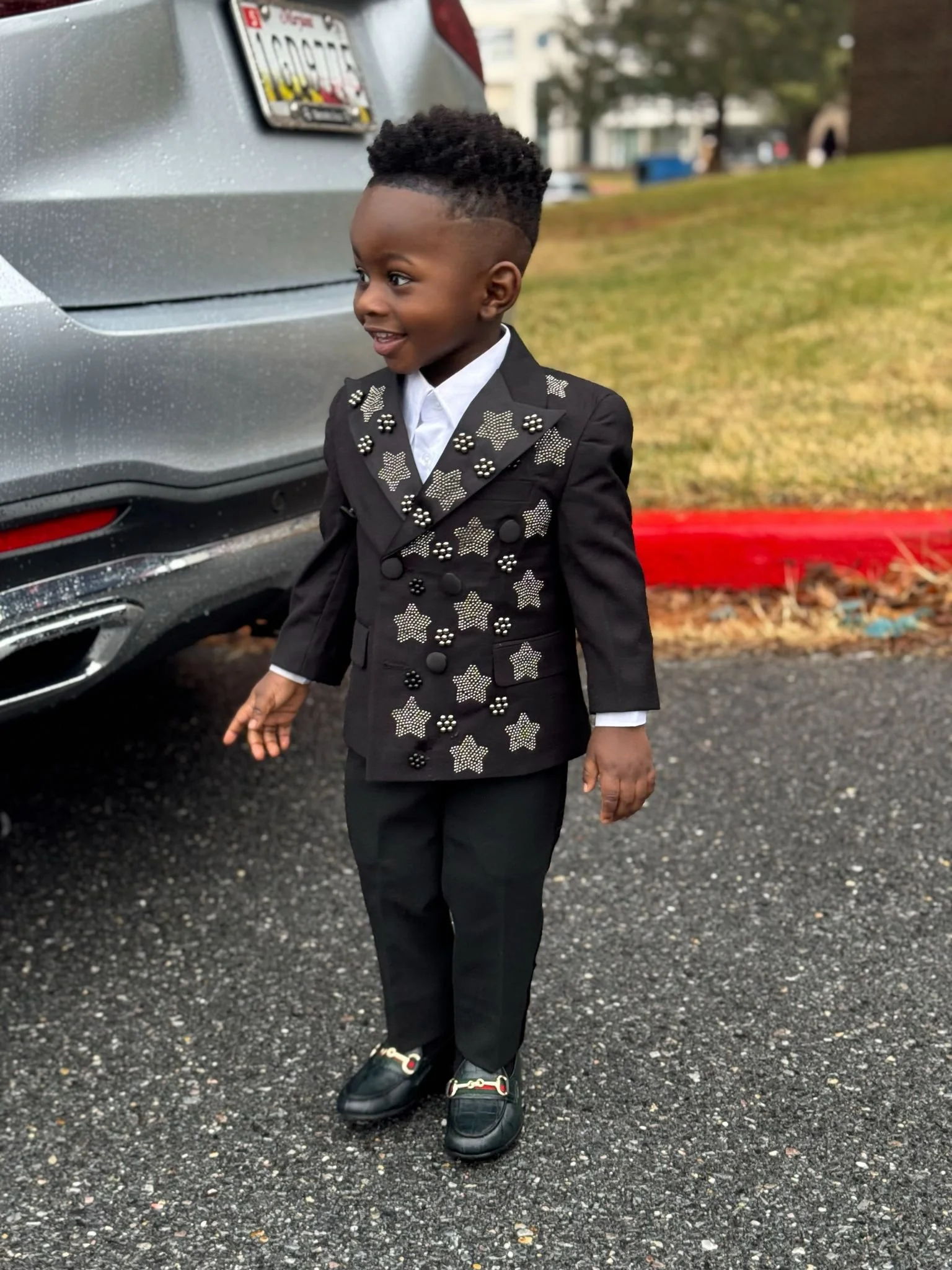 Young boy dressed in a black suit with star patterns and embellishments, standing outdoors next to a grey car with a rain-dotted surface.