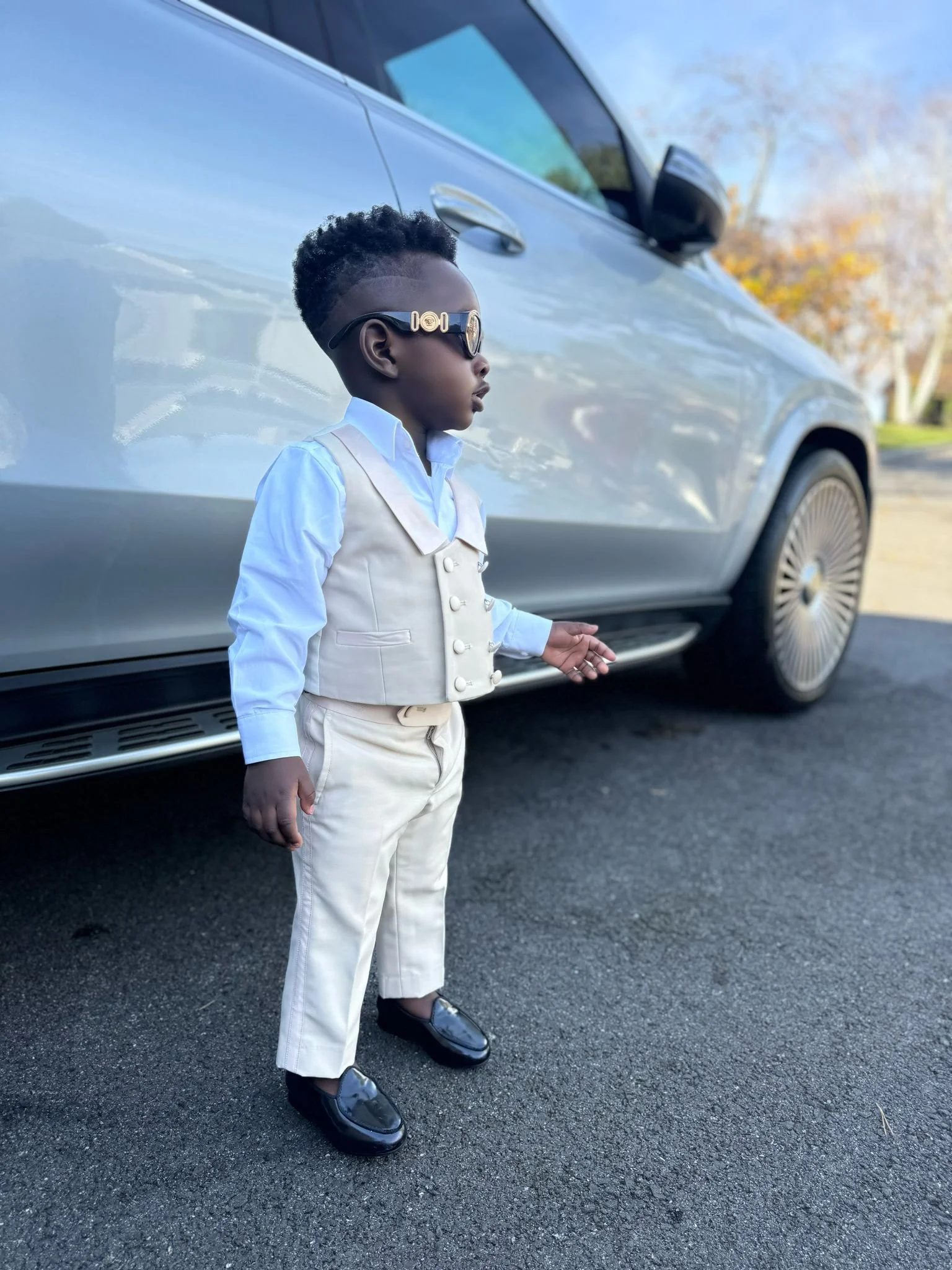 Young boy dressed in a cream vest, cream trousers, light blue shirt and sunglasses, standing on a paved surface beside a silver luxury car with trees in the background.