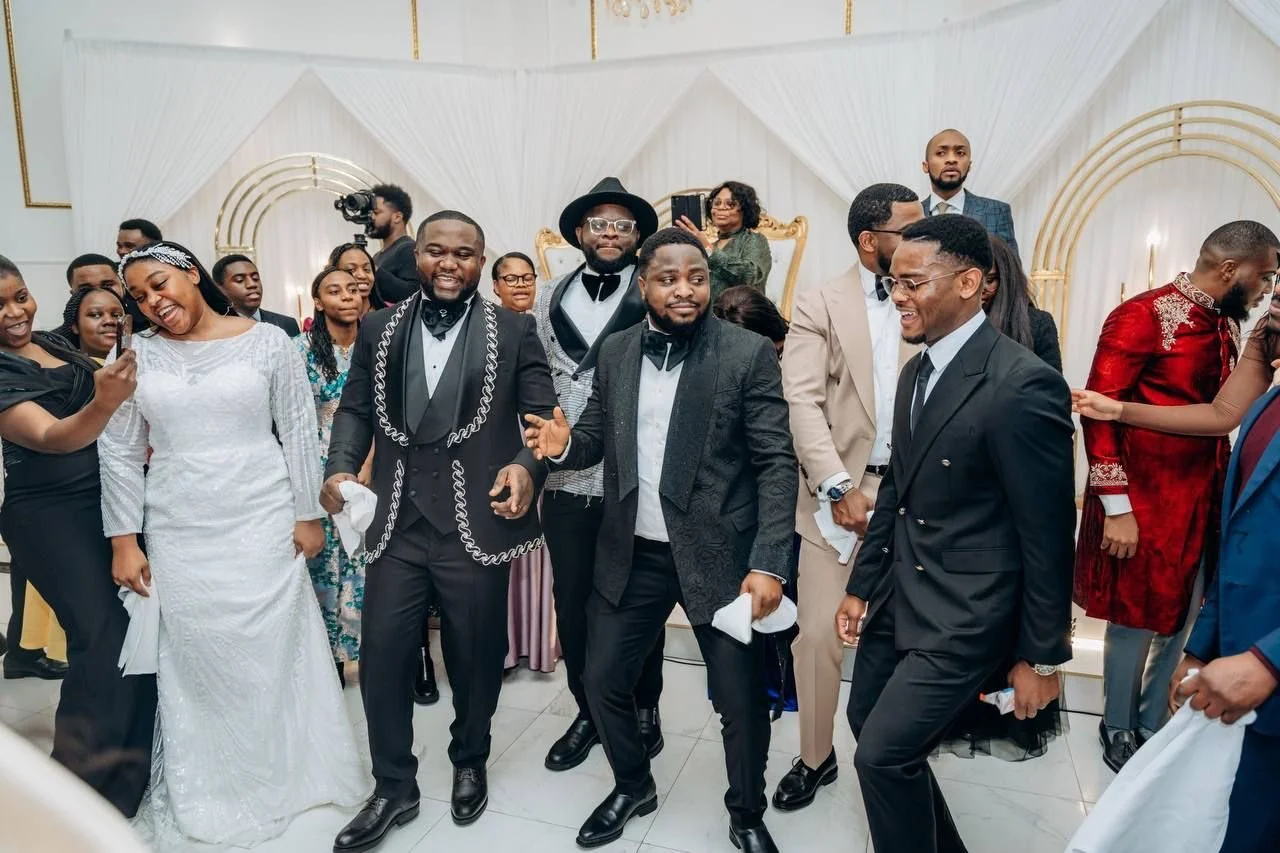 Group of people dancing and celebrating at a wedding reception, some dressed in formal attire, with a bride in a white gown smiling in the foreground.