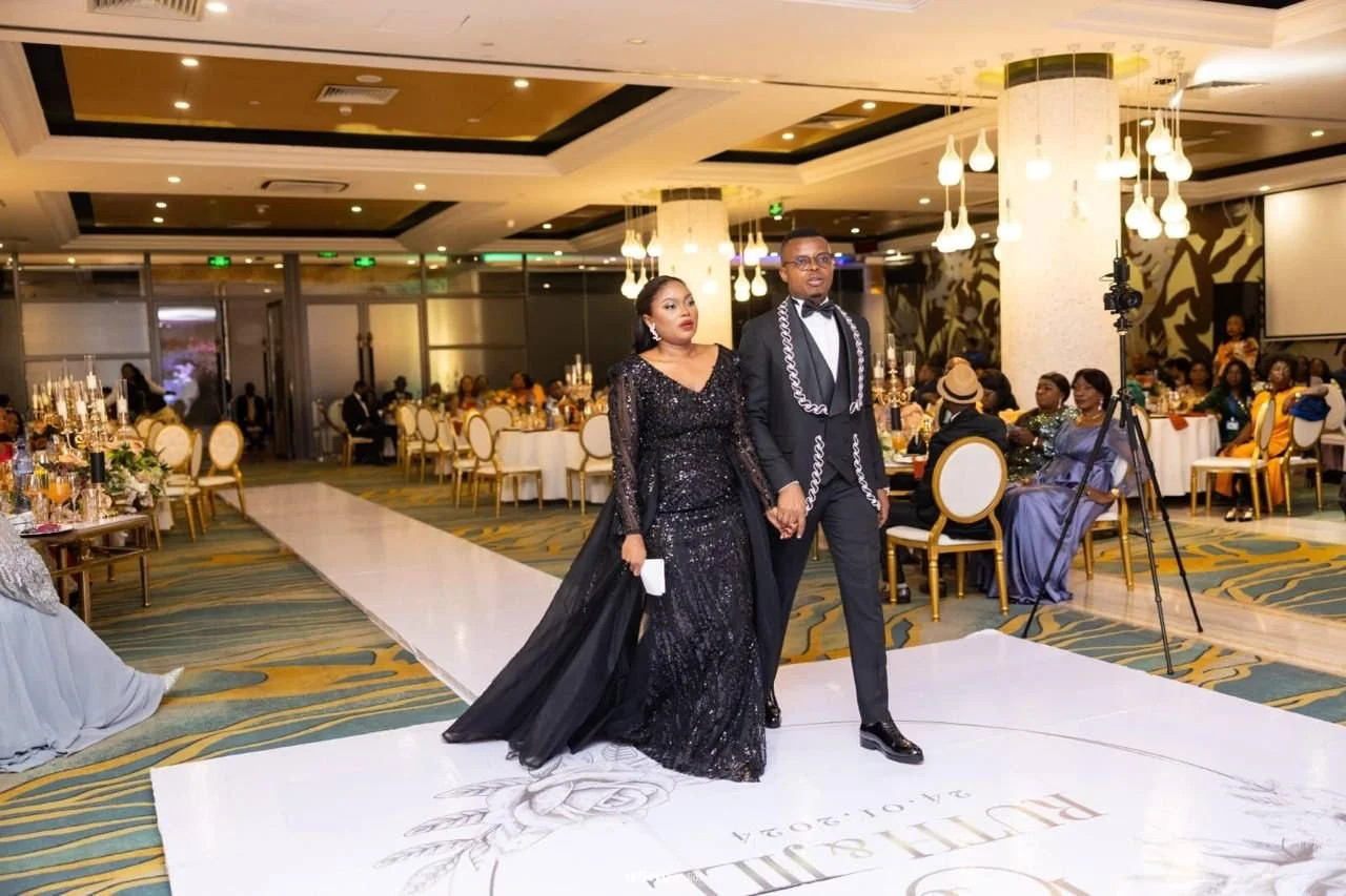 A couple dressed in formal attire walking hand in hand on a wedding dance floor during a celebration, with guests seated at decorated tables in the background.