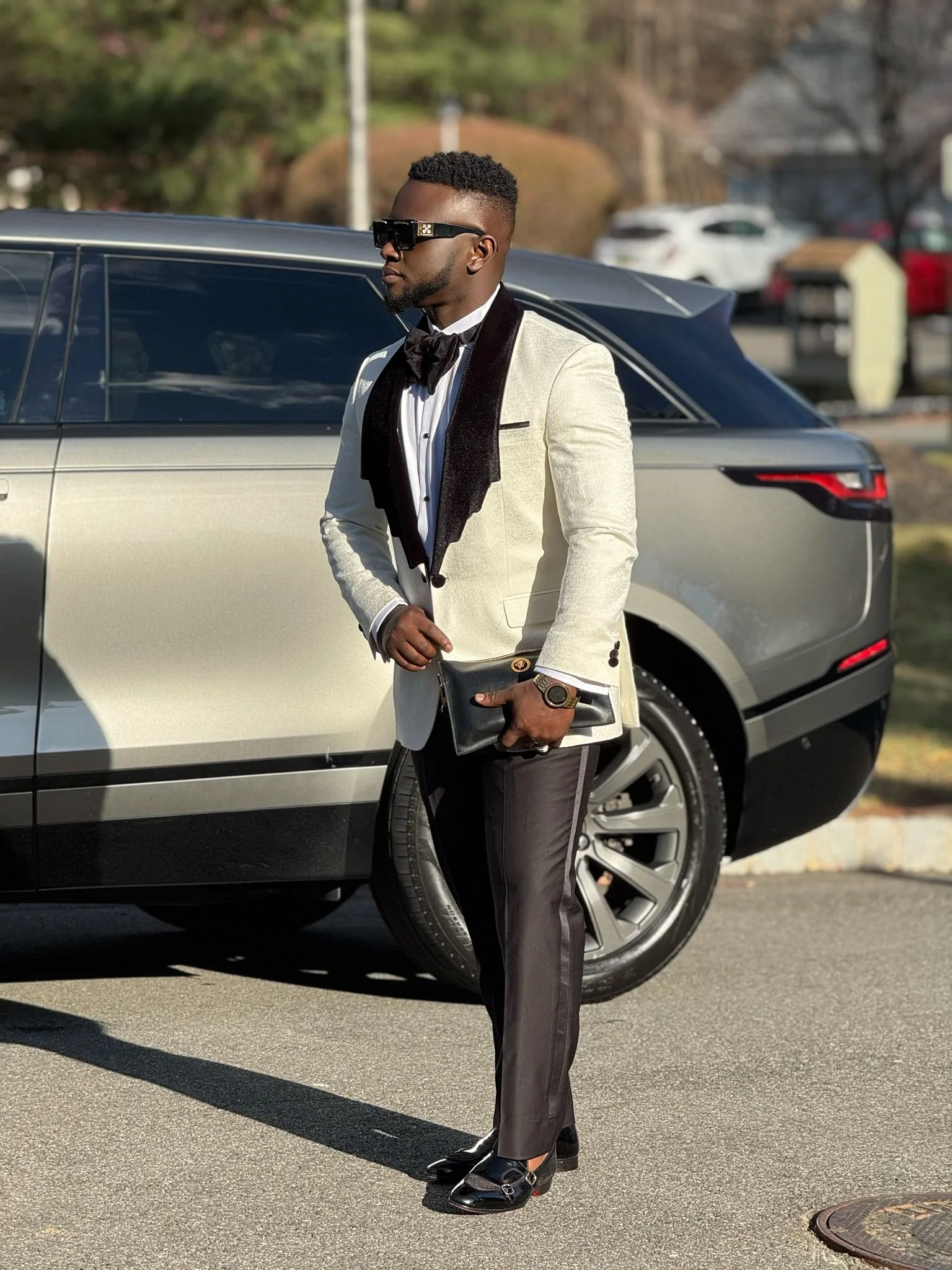 A stylish man in a white blazer, black pants, and black shoes stands in front of a silver car. He is wearing sunglasses, a black bow tie, and holding a black clutch.