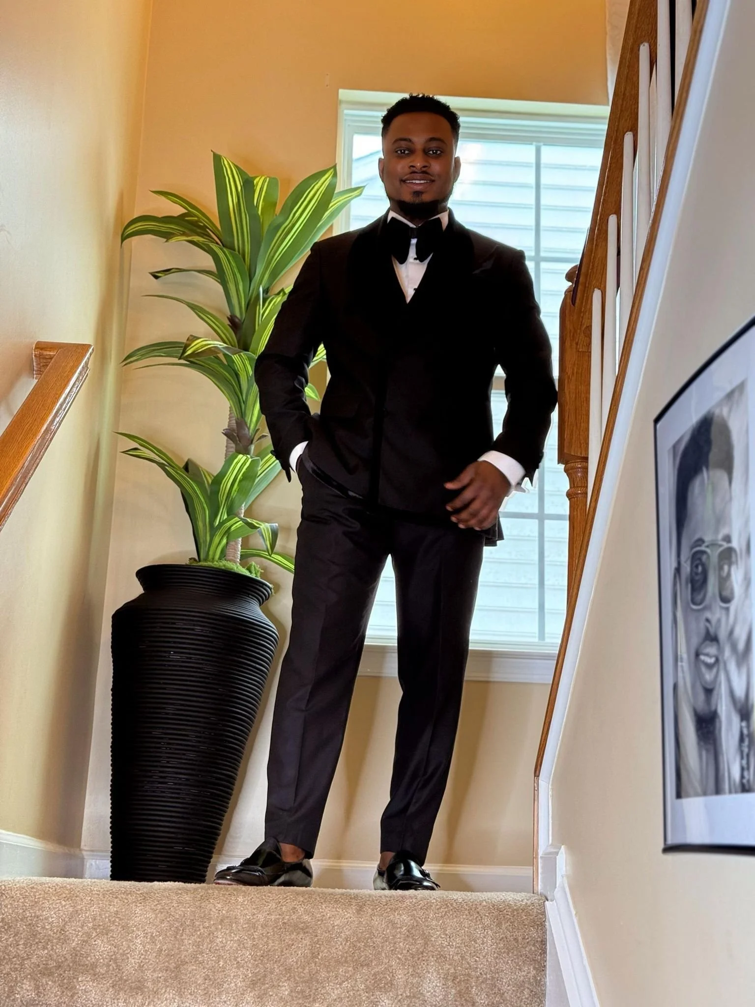 Man in black tuxedo and bow tie standing on staircase next to large potted plant and framed picture, with a window behind.