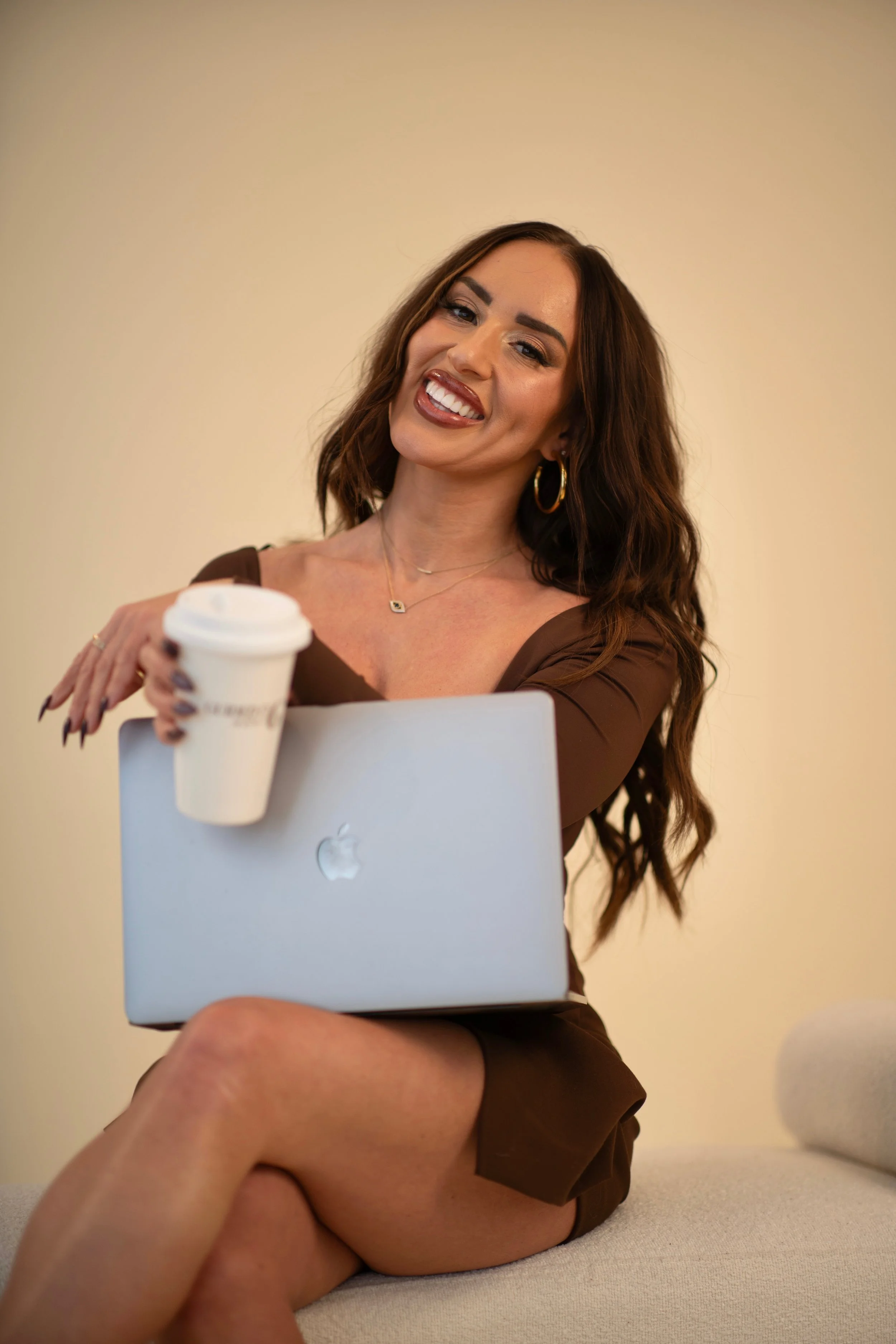 A woman with long brown hair, wearing gold hoop earrings and a brown top, sitting cross-legged on a light-colored surface, holding a white disposable coffee cup, smiling and looking at the camera with an Apple computer in front of her, set against a plain, beige background.