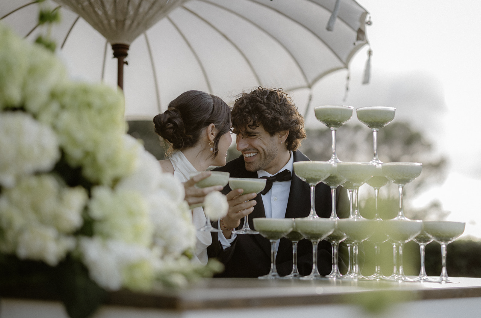 A couple at a wedding celebration sharing a toast with matcha champagne in coupe glasses, under a white umbrella, with a matcha champagne tower and floral decoration in the foreground.