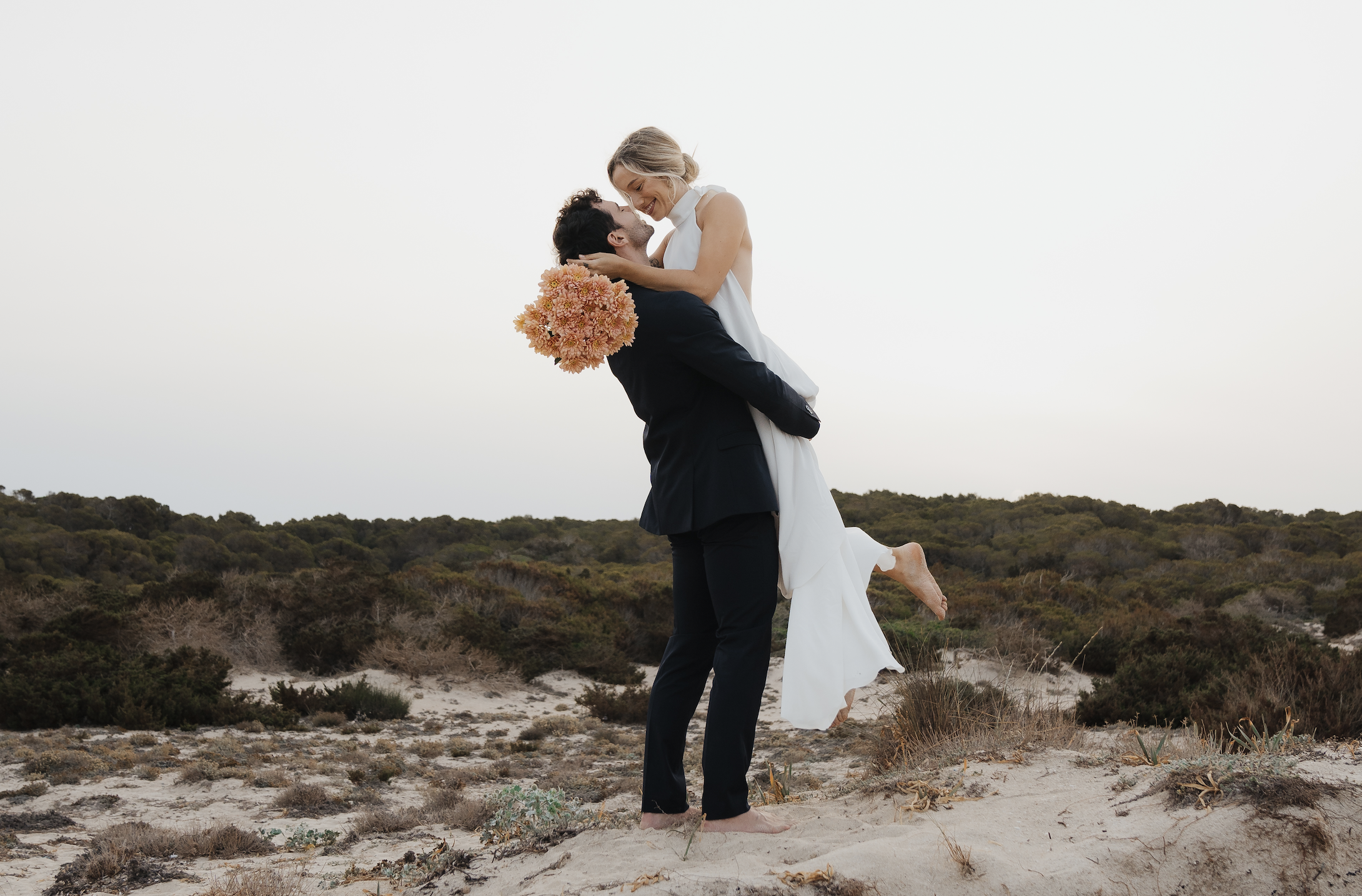 A couple in wedding attire, with the man lifting the woman, on a sandy area with bushes and trees in the background.