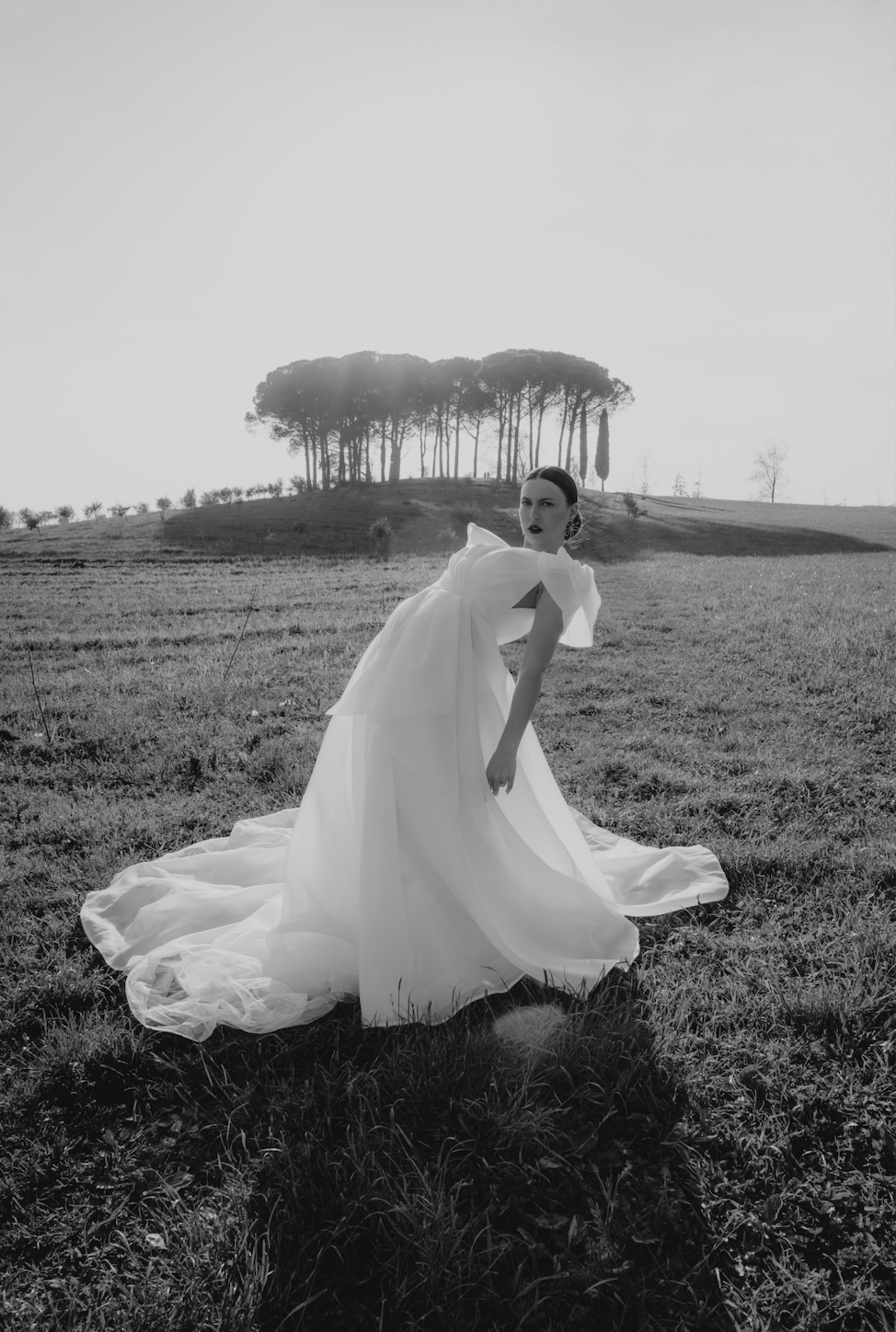 Black and white photo of a woman in a long, flowing white dress standing on grass in a field with rolling hills and a group of tall trees in the background.