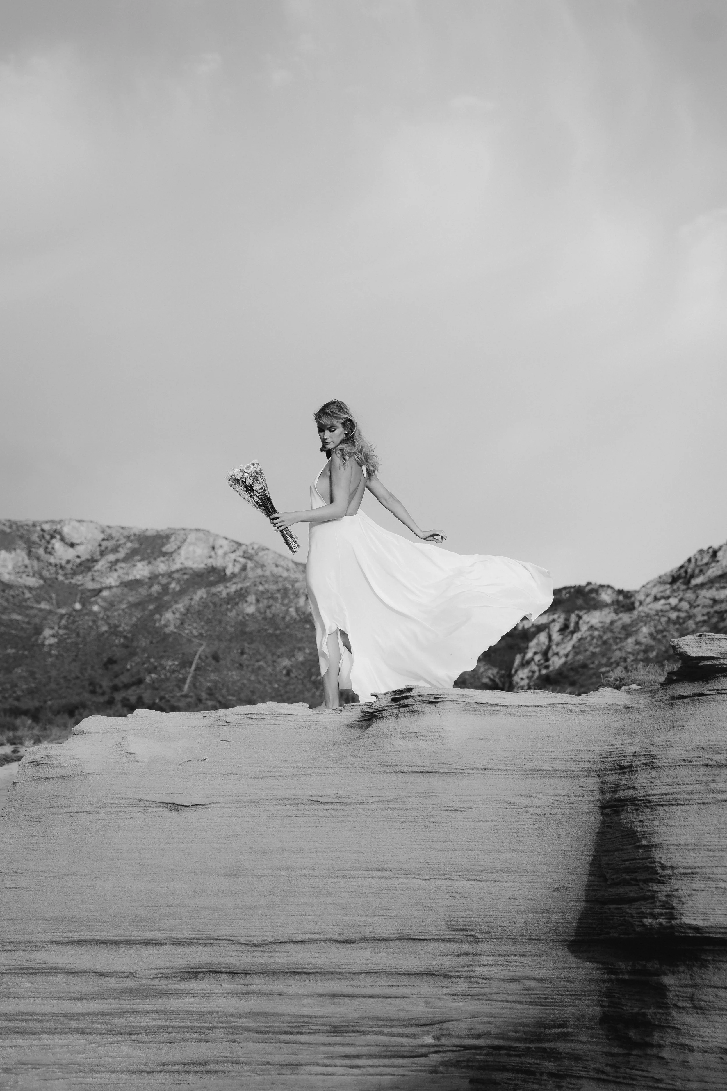 Black and white photo of a woman in a flowing white dress holding a bouquet of flowers, standing on a large piece of driftwood in an outdoor landscape with mountains in the background.