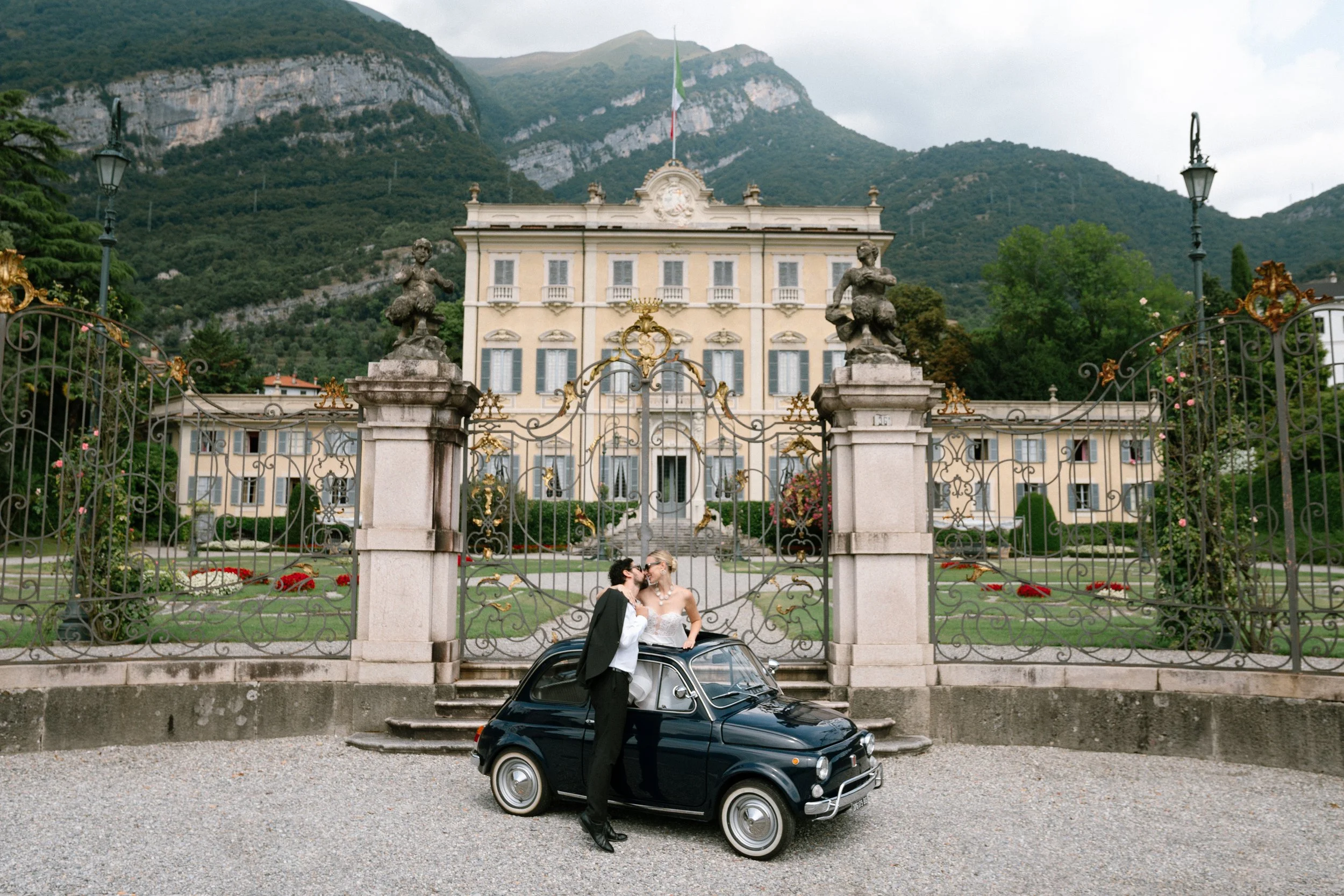 A couple dressed in wedding attire sharing a kiss while standing on a vintage black car in front of a large ornate building with a gated entrance, surrounded by greenery and mountains.