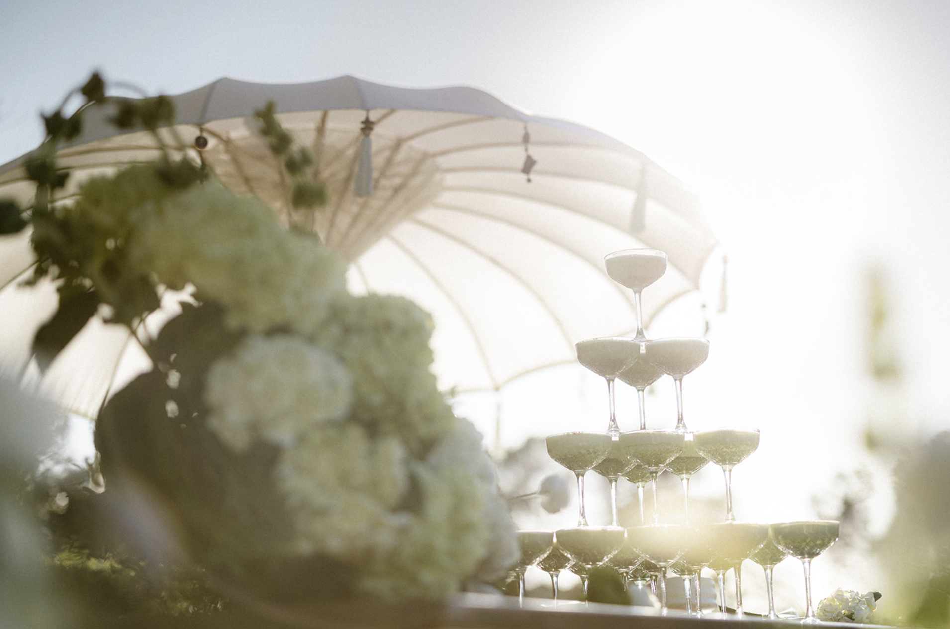 A matcha latte champagne tower with multiple glasses stacked in a pyramid shape, with an umbrella and flowers in the background, outdoors during daytime.
