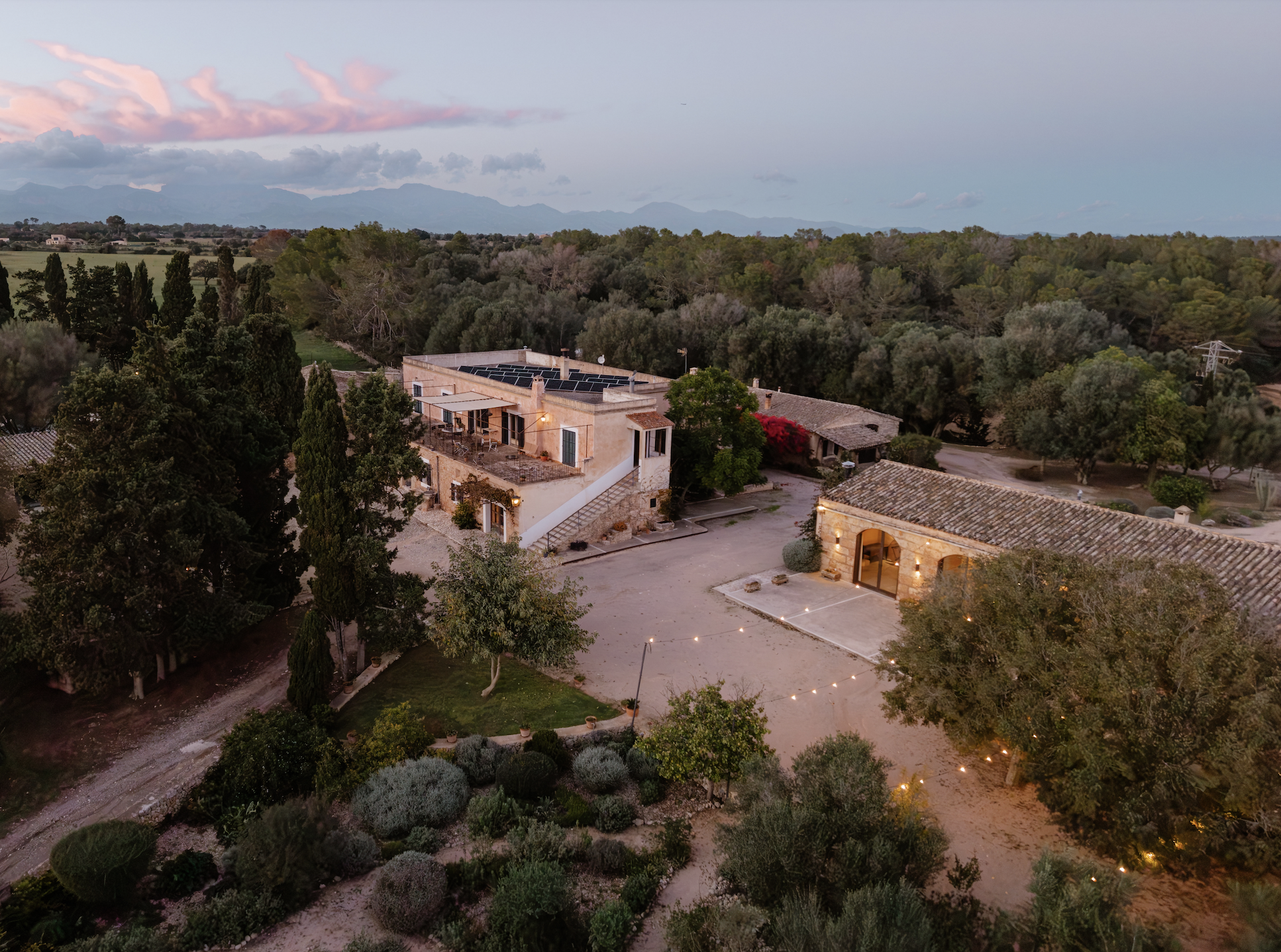Aerial view of a countryside estate at dusk, featuring a main building with solar panels on the roof, surrounded by trees, greenery, and outdoor string lights.