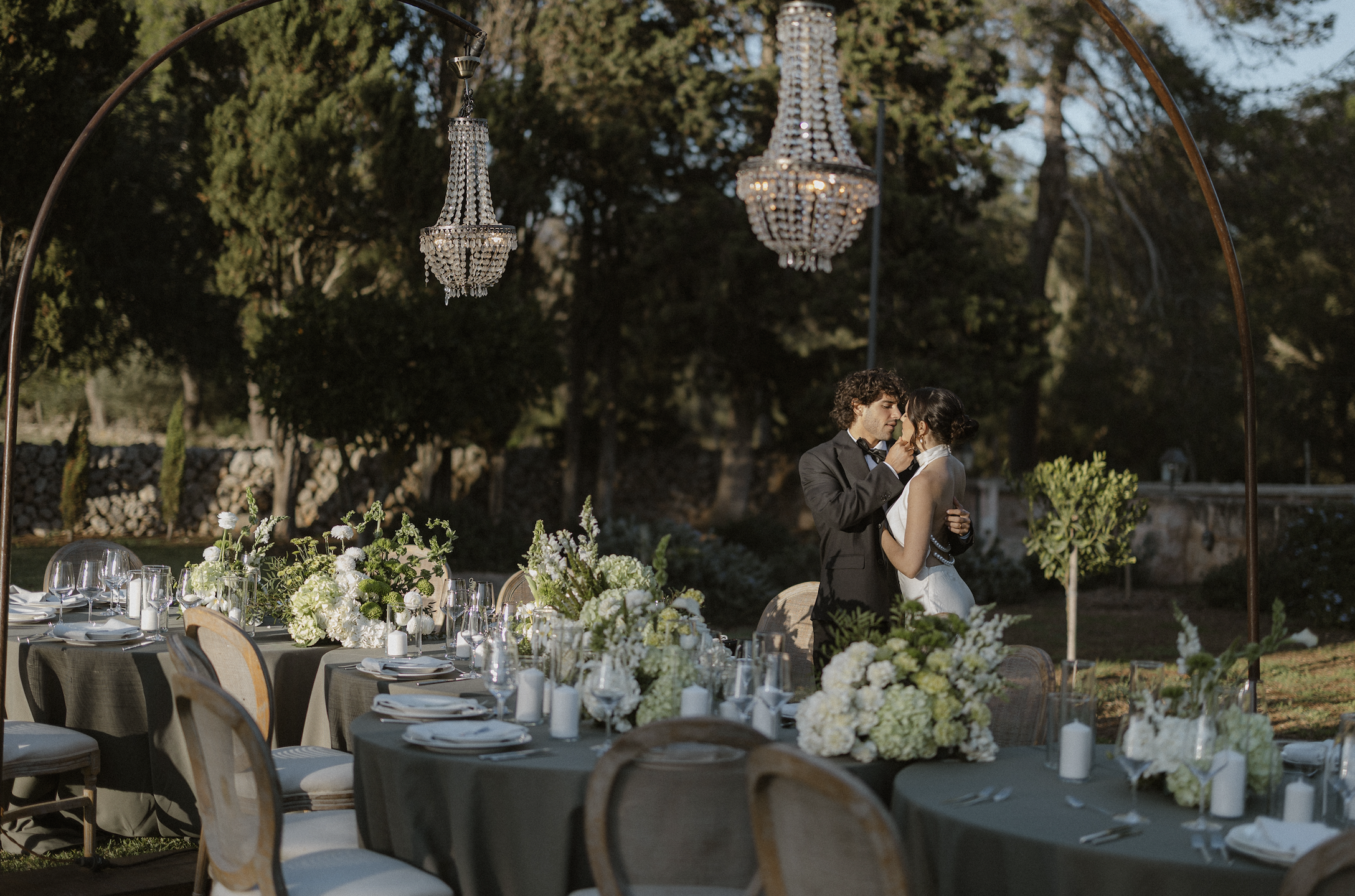 A bride and groom share a romantic moment at their outdoor wedding reception, under a decorative arch, with elegant table settings and floral arrangements.