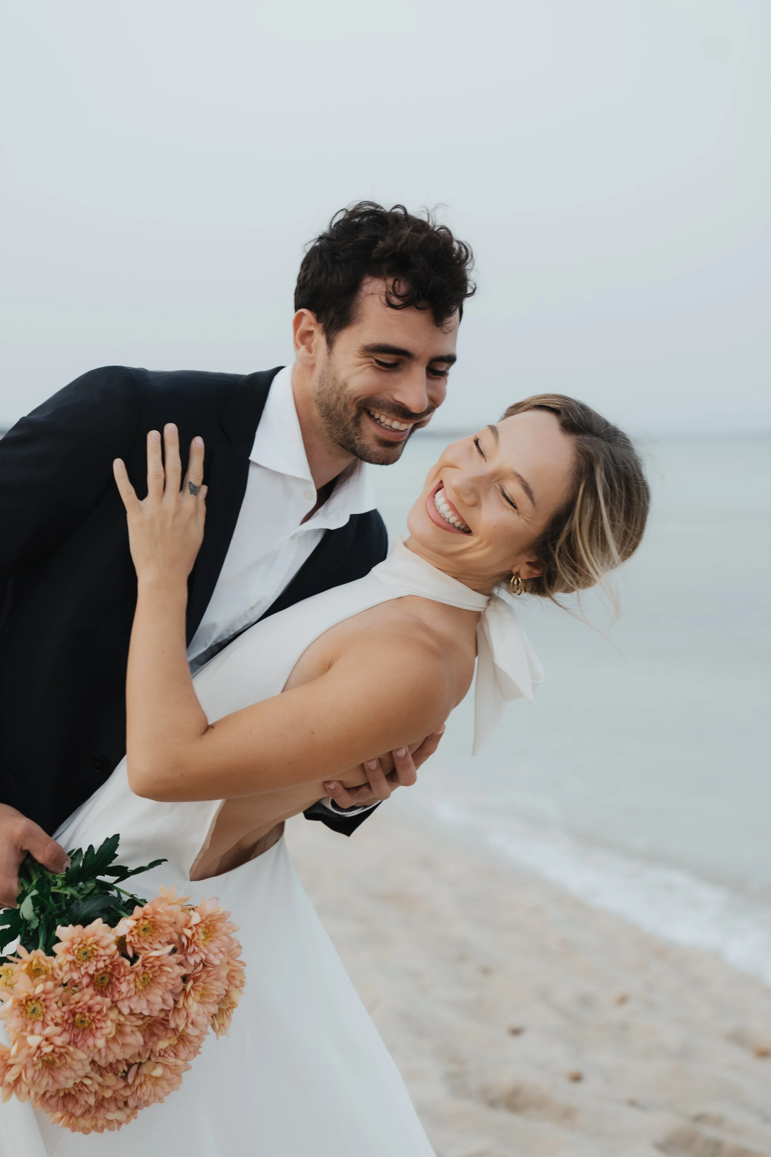 A happy couple in wedding attire, the groom holding the bride, laughing on a beach.