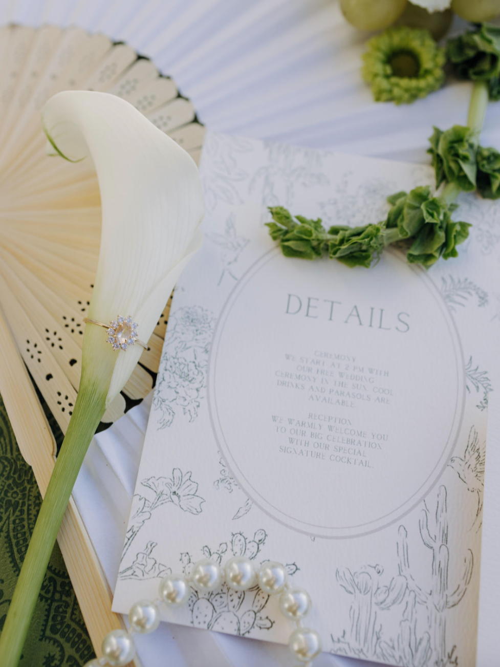 Wedding invitation, white calla lily, green floral necklace, pearl bracelet, and decorative paper fan.