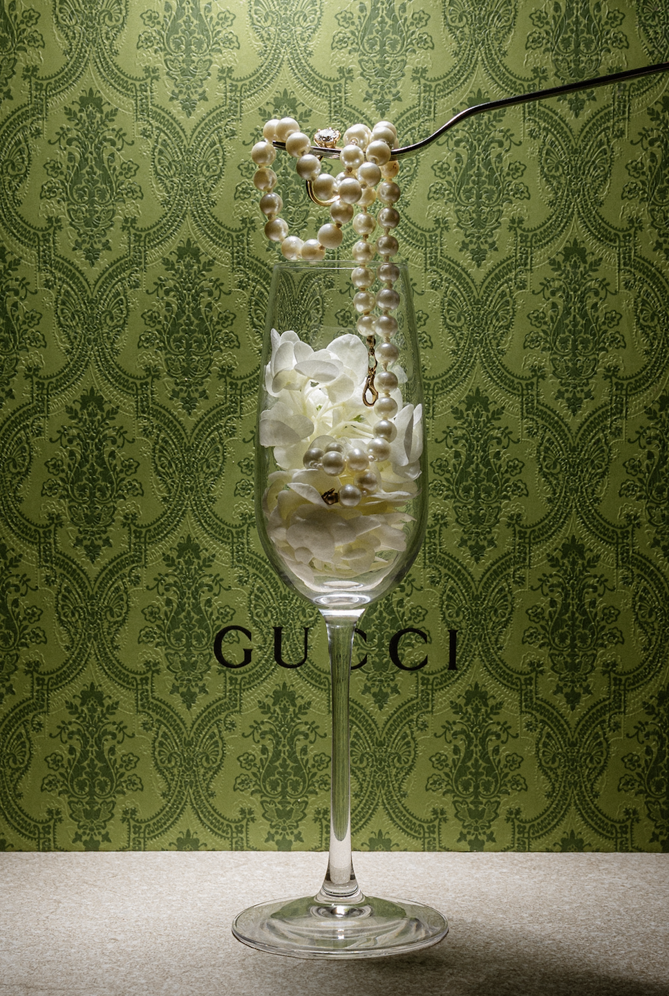 A tall champagne flute filled with white flower petals, set against a green patterned Gucci backdrop. Floating pearl necklaces and rings are placed inside and on top of the glass.