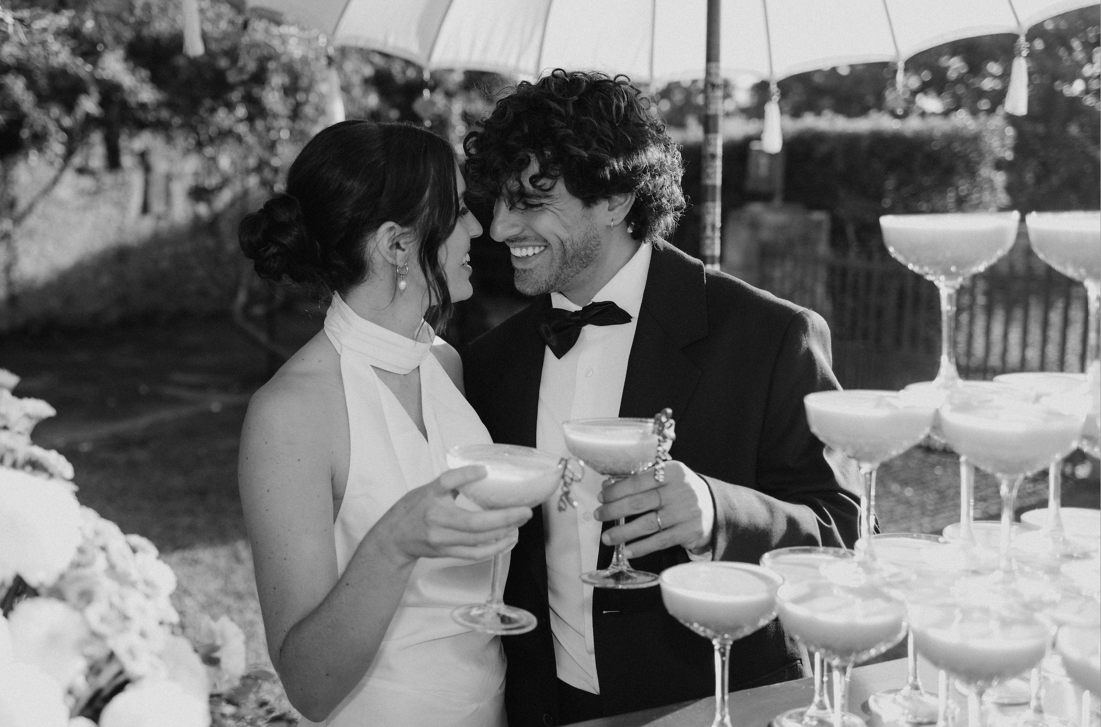A black-and-white photo of a bride and groom smiling and holding drinks at their outdoor wedding reception, with large umbrellas overhead and a table with champagne glasses.