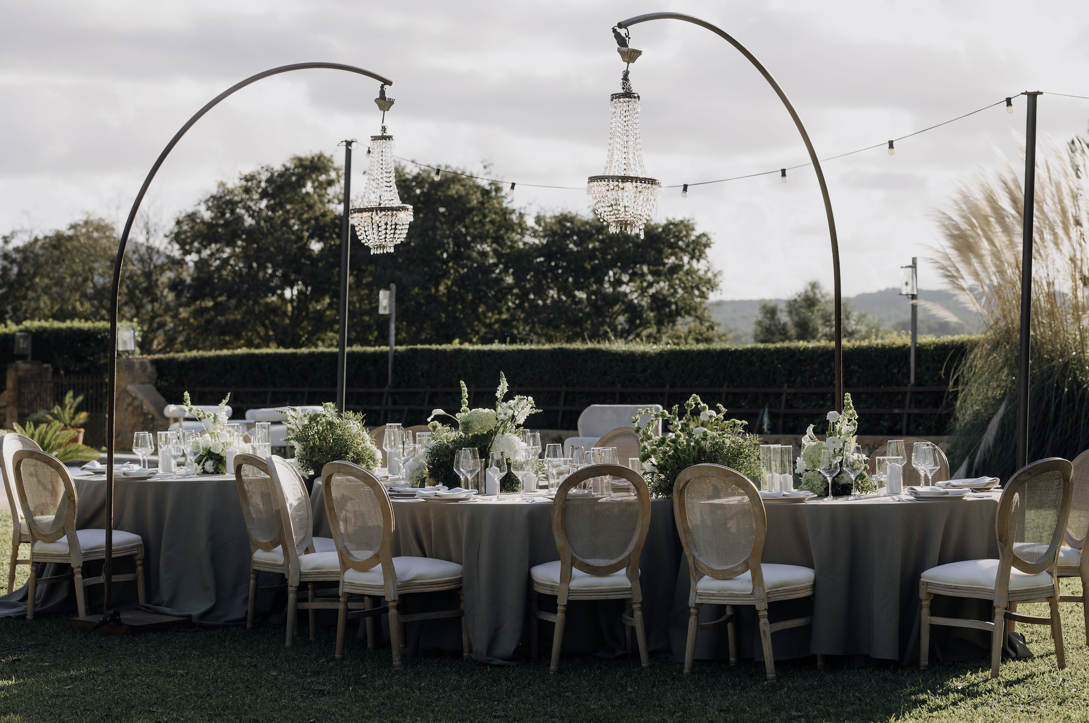 Outdoor banquet table set with floral centerpieces, glassware, and tableware during daytime, with hanging chandeliers and string lights, greenery and trees in the background.