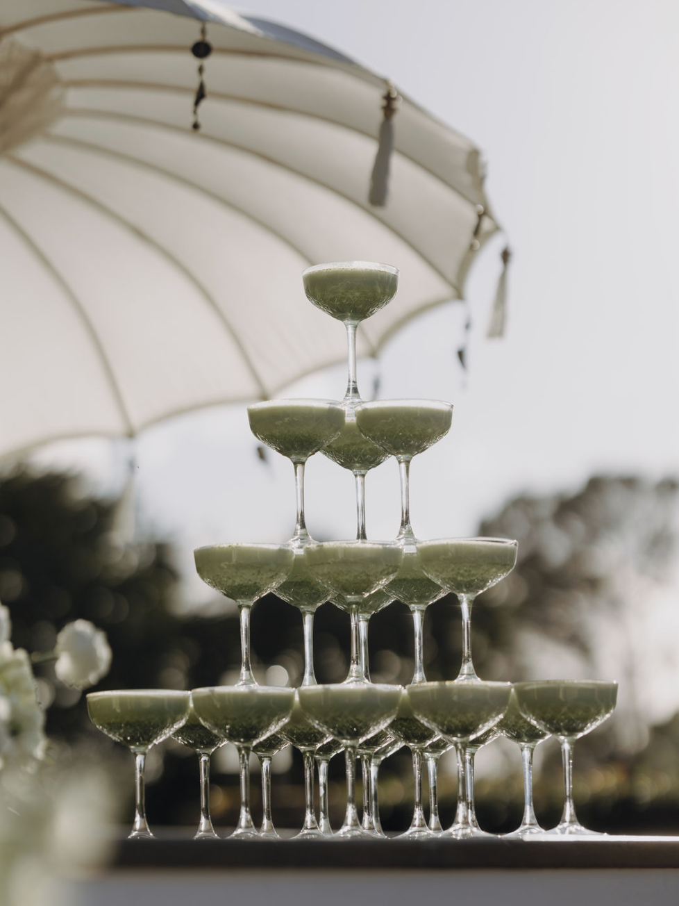 A champagne tower made of several layers of green-colored cocktail glasses arranged in a pyramid, with a large white umbrella in the background.