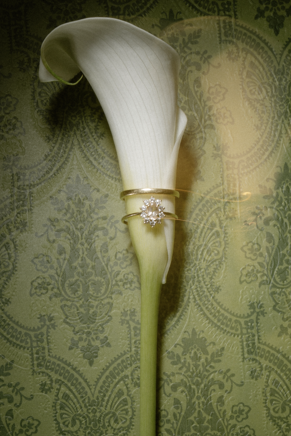 A white calla lily flower with two rings, one with a diamond flower design, placed on its stem, against a patterned green background.