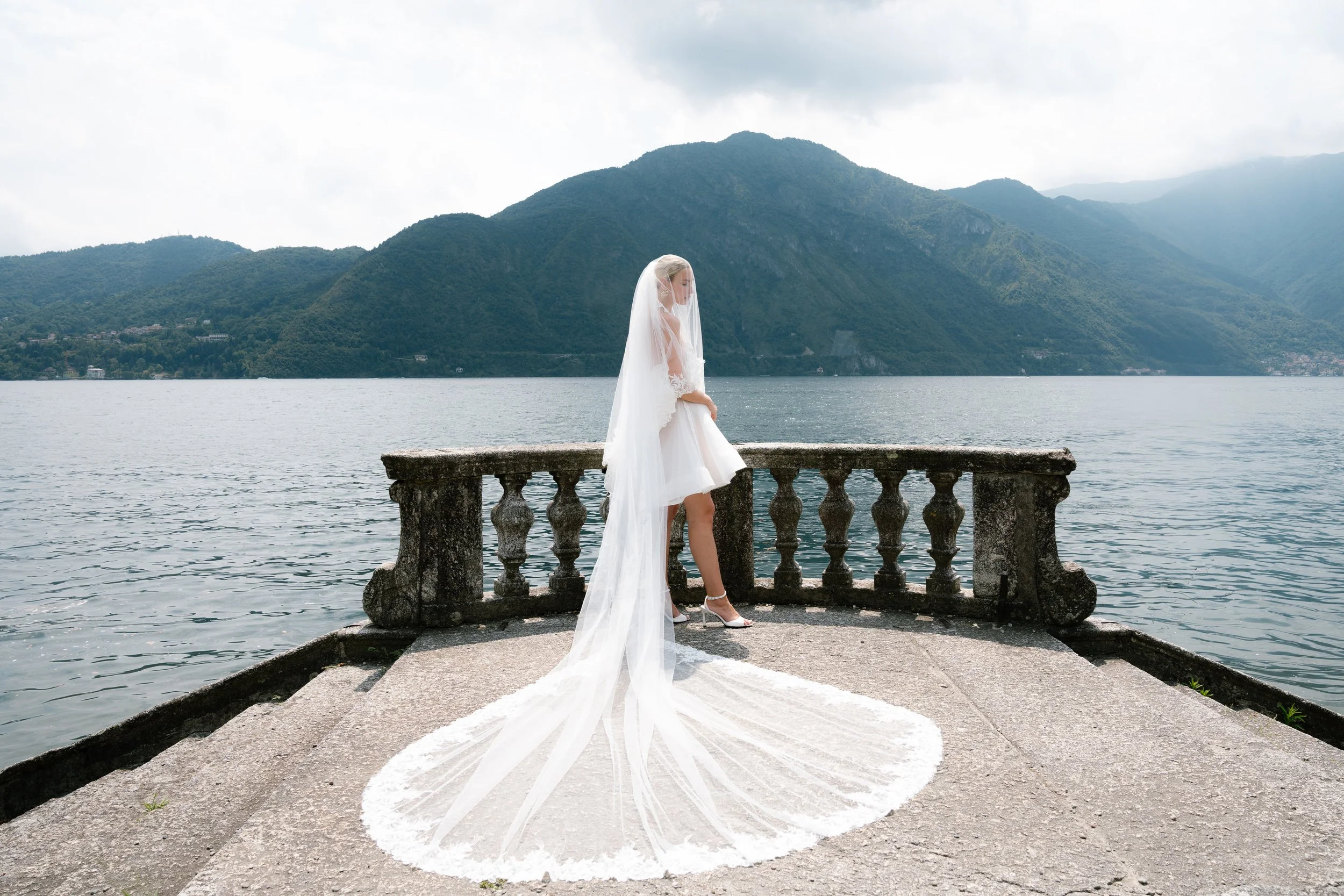 A bride in a white wedding dress and veil stands on a stone pier with a lake and mountains in the background.