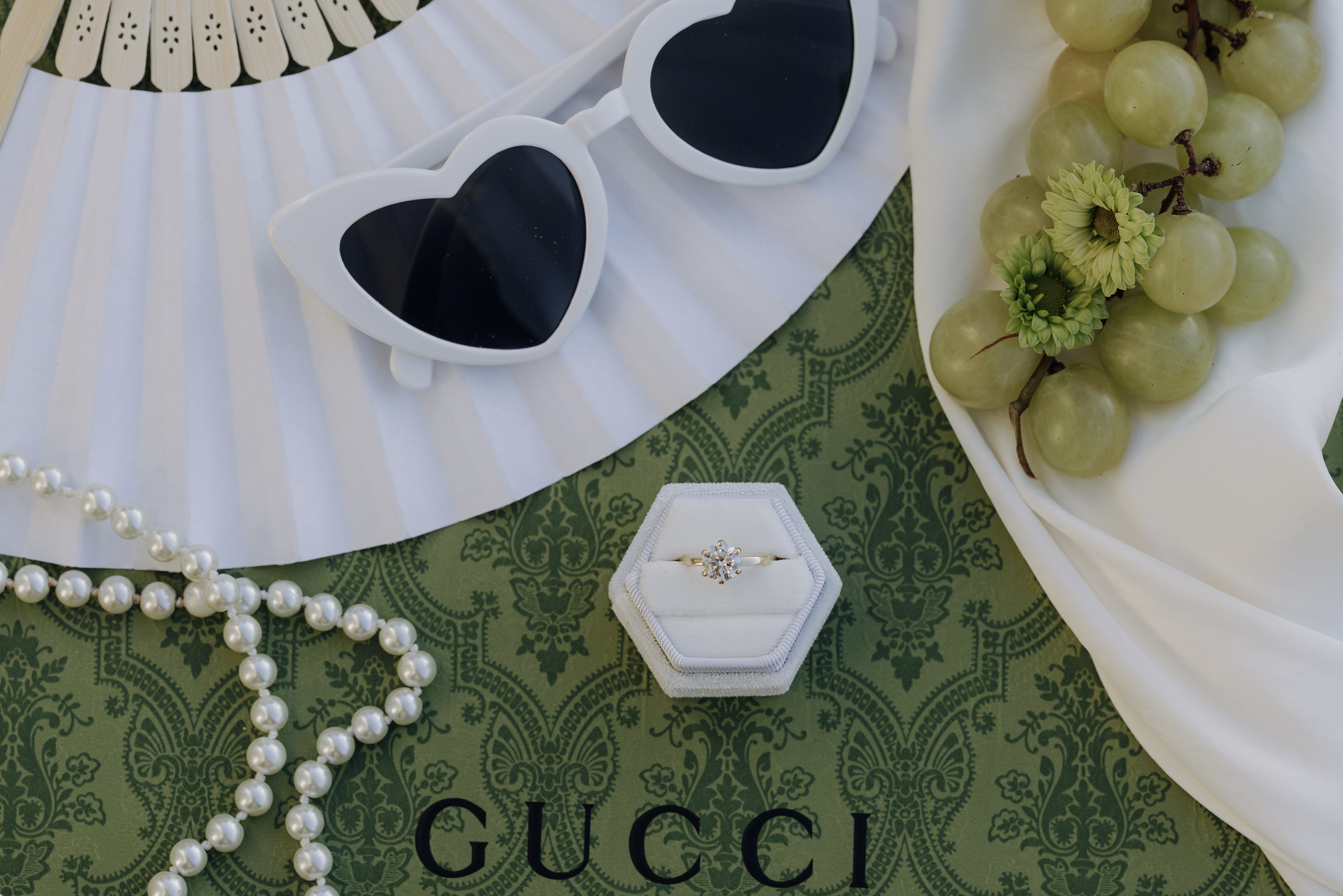 A display of a pearl necklace, heart-shaped sunglasses, a diamond ring in a white ring box, green grapes with small flowers, and a white fan on a green patterned fabric with part of a white cloth, with the Gucci logo on the fabric.