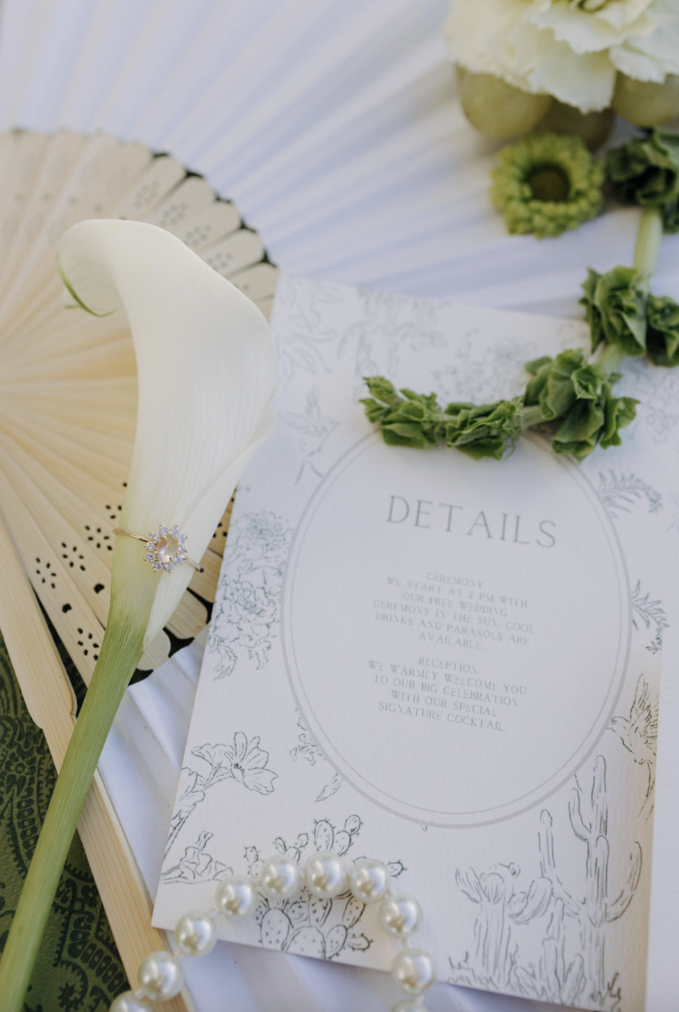 Wedding invitation surrounded by a calla lily, pearl necklace, green floral crown, and a wooden fan, with a white flower bouquet in the background.