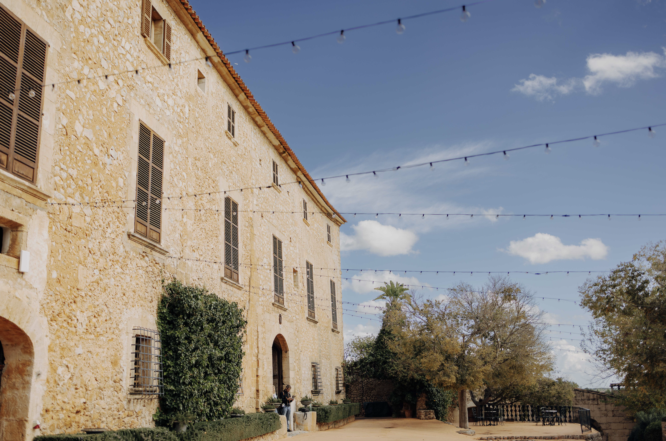 A stone building with multiple windows and shutters, greenery, trees, outdoor seating, and string lights under a blue sky with clouds.