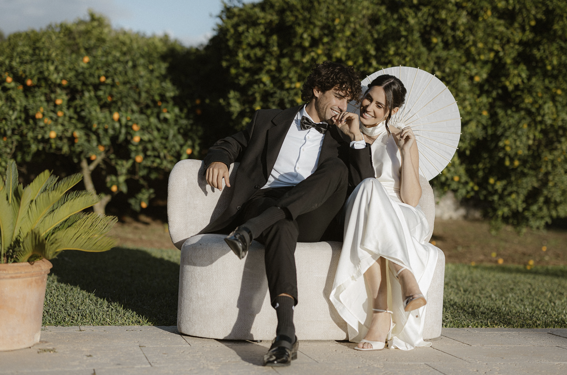 A smiling couple sitting on a beige couch outdoors, dressed in formal attire, with a woman holding a white parasol, surrounded by greenery and orange trees.