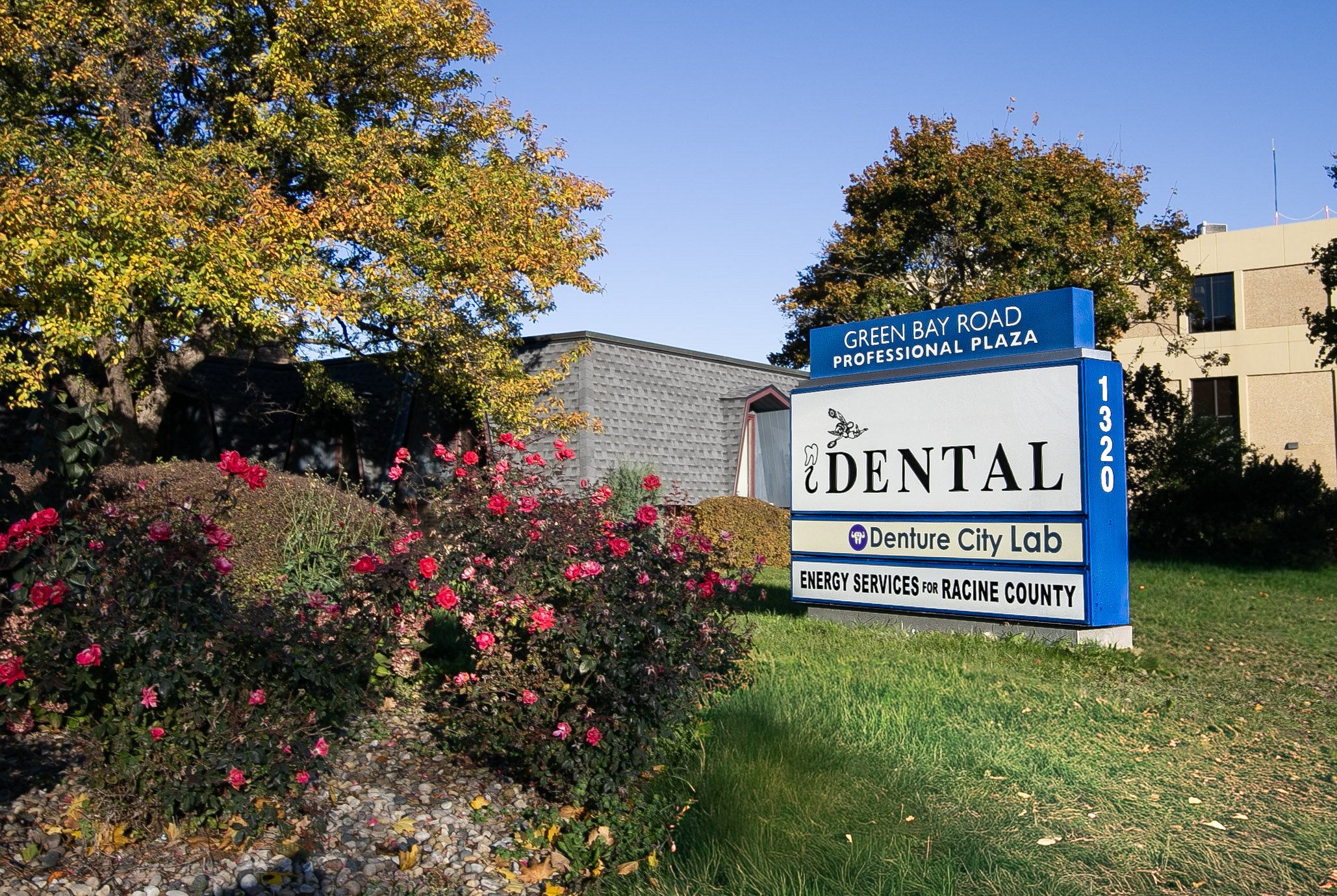 Sign for Green Bay Road Professional Plaza with dental office, Denture City Lab, and Energy Services for Racine County. Surrounded by trees and pink flowers on a sunny day.