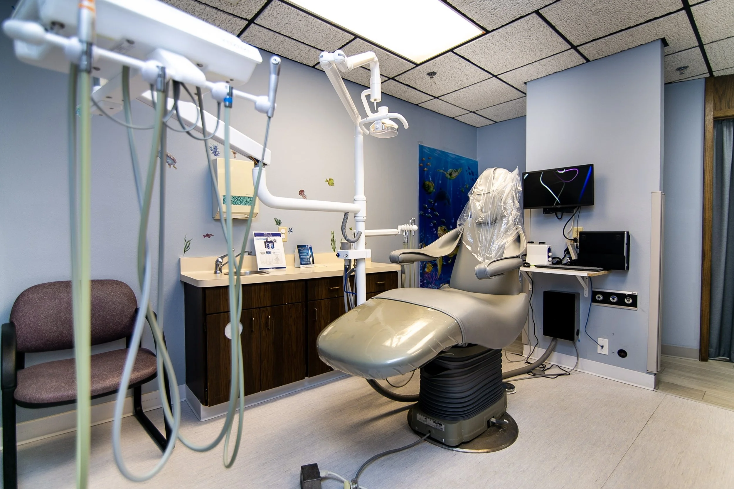 An empty dental examination room with a dental chair, dental light, and medical equipment, including a monitor on the wall.