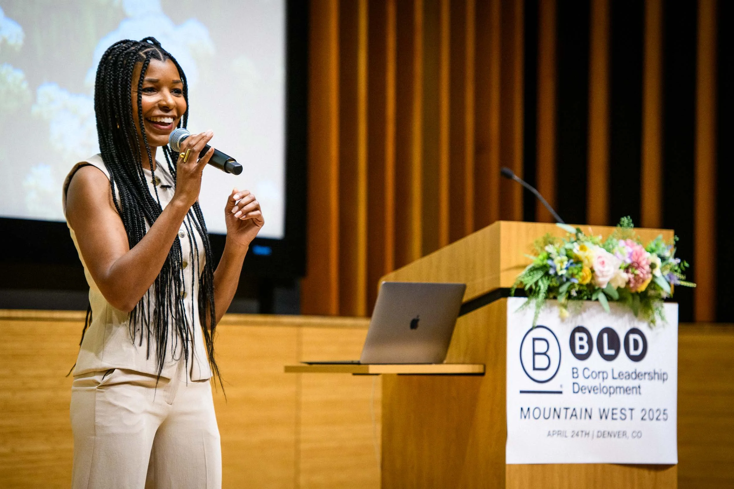 A woman with braided hair smiling while speaking into a microphone at a conference or event, with a podium and large screen behind her displaying a presentation slide, and a laptop on the table next to a floral arrangement.
